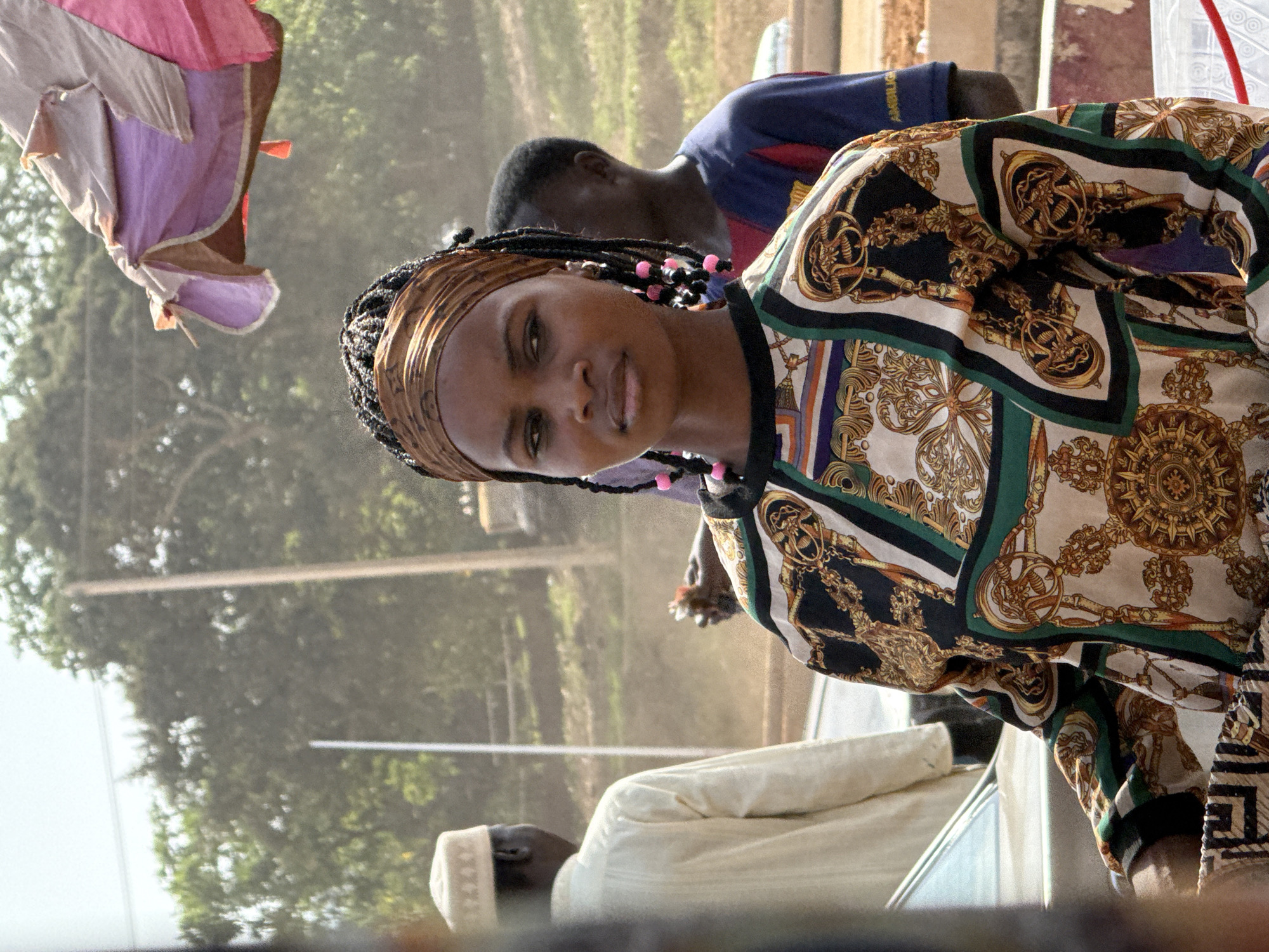 A medium shot, taken from a slightly high angle, shows a woman in a busy outdoor setting. The woman, positioned in the foreground, is wearing a patterned blouse and a head wrap. She has dark skin, black braided hair with colorful pink beads, and is looking directly at the camera.
Behind her and slightly out of focus are other people, including a man wearing a blue shirt, and another wearing a white robe and cap.
The background appears to be a bustling outdoor location, possibly a market or transportation hub, with trees and other vague structural elements.
Based on the prompt, the location is in Abuja, Nigeria, however, there is no specific detail visible in the image that confirms this location.