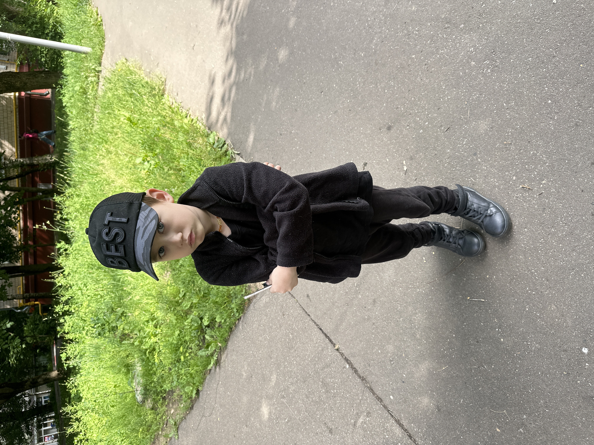 A young boy stands on an asphalt path next to a grassy area. He is wearing a black fleece jacket, black pants, black baseball cap with the word "BEST" printed in grey letters, and black boots. He is holding a small silver object in his right hand. The boy is looking towards the camera with a neutral expression. The background shows a residential building with red and yellow accents, trees, and green grass. The scene appears to be outdoors on a sunny day. The lighting suggests it is daytime. The presence of residential buildings and lush greenery hints at a typical urban park or courtyard setting within Moscow, Russia.