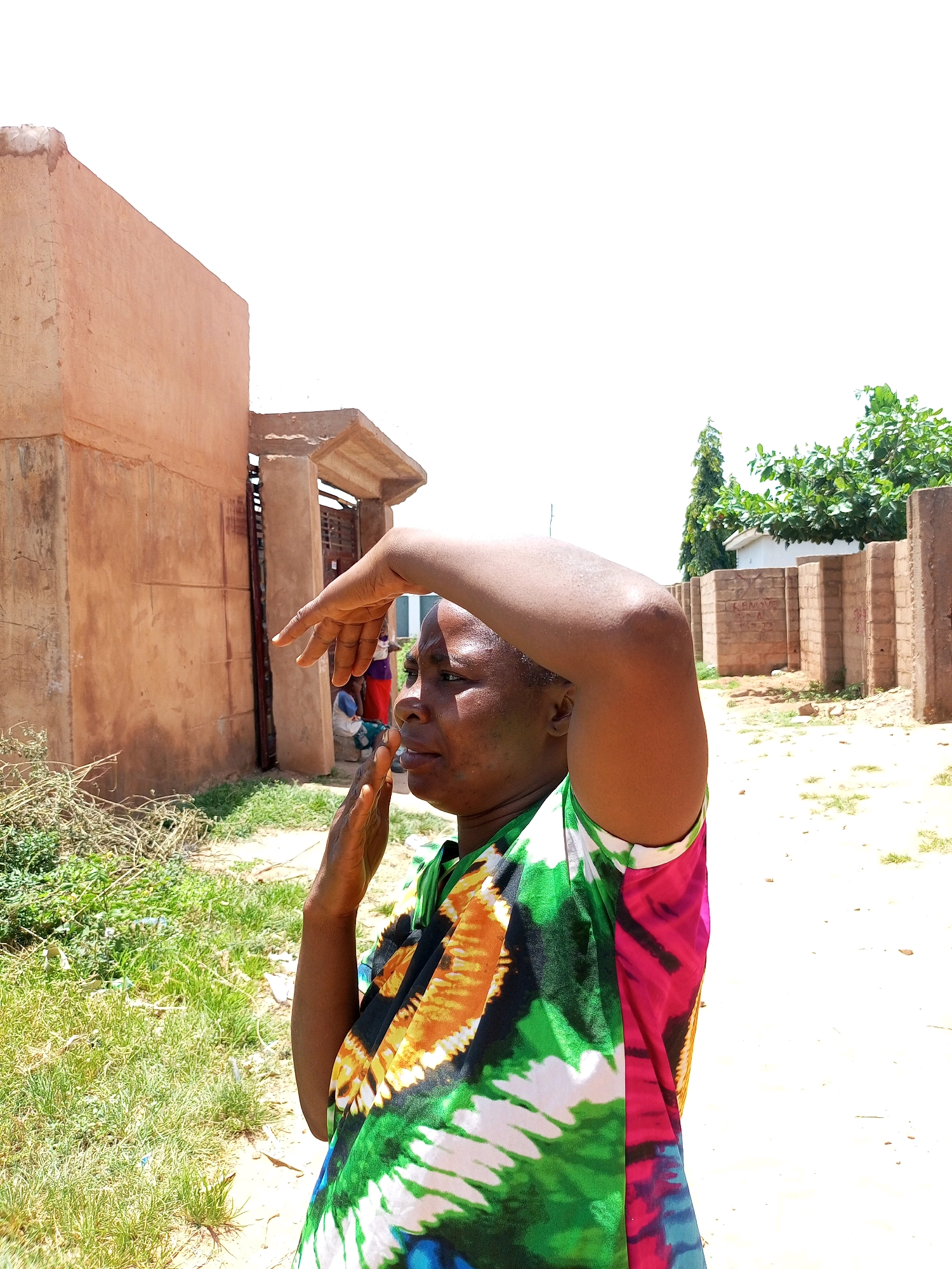The image shows a woman standing outdoors in what appears to be an unpaved area in Tempure, Nigeria. She is seen from the side, looking upwards with her right arm raised and hand near her forehead, possibly shielding her eyes from the sun. She is wearing a colorful, tie-dye patterned shirt with green, yellow, orange, and pink hues.
In the background, to the left, is a building with an unfinished, earthy-toned facade and a covered entryway. To the right, there is a wall constructed from reddish-brown bricks, and beyond that, a tree with dense green foliage and another building with a white exterior. The ground is composed of light-colored dirt and sparse vegetation. The sky is bright and overcast.