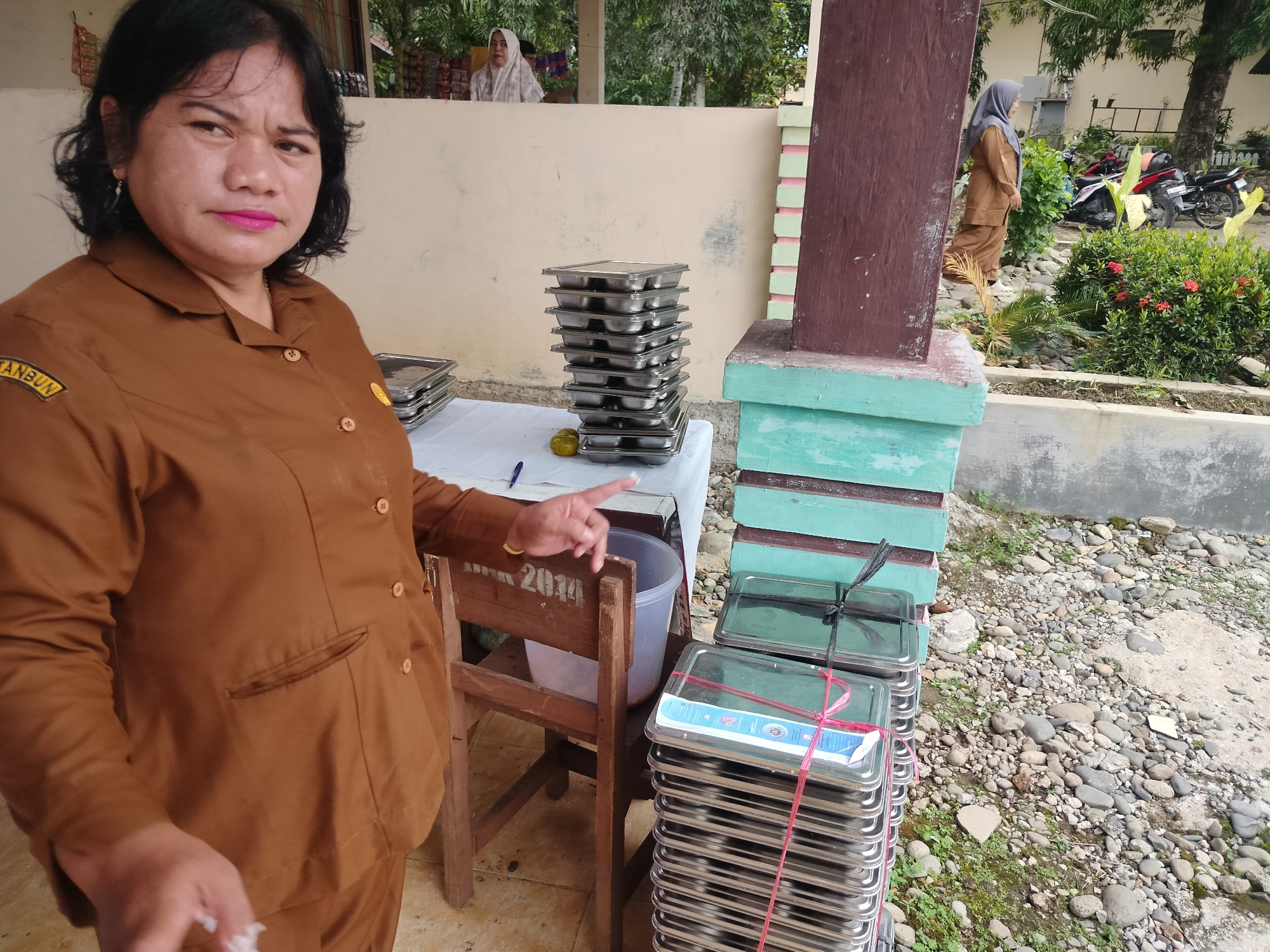 In Purwodadi, Indonesia, a woman wearing a brown uniform is standing near stacks of metal trays. She is in the foreground, facing the camera with a slight smile and pointing to the right. Behind her, on a table covered with a white cloth, are more trays. In the background, two other women are visible. One is wearing a hijab and a brown outfit, standing behind a wall. Another woman, also in a hijab and brown outfit, is walking away from the camera to the right. The environment appears to be outdoors, with a wall, trees, bushes, and a motorcycle in the background. The ground is covered with small stones and some greenery.