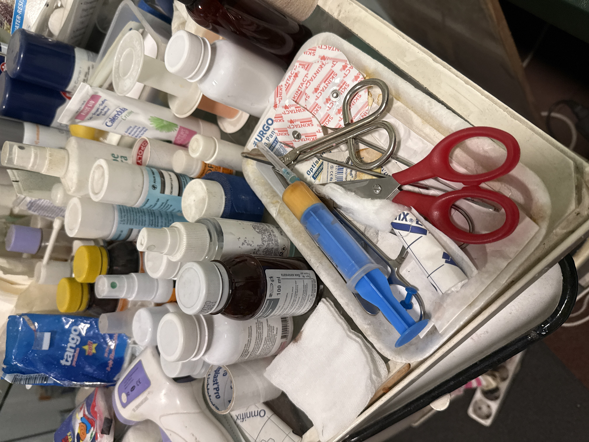 The image shows a cluttered tray containing various medical supplies. The tray appears to be part of a larger medical cart. Visible items include a large blue syringe, scissors with red handles, cotton pads, and multiple bottles and tubes of medications and creams, including "Calendula Cream" and "Tango" ointment. Some of the bottles have labels in Cyrillic script. Brands such as "Omnifix", "Plast Pro", "Optisk", "Urgo", and "Skintact" are visible. Based on the Cyrillic script and the brand "Optisk" (a Bulgarian brand), the location is likely Sofia, Bulgaria.