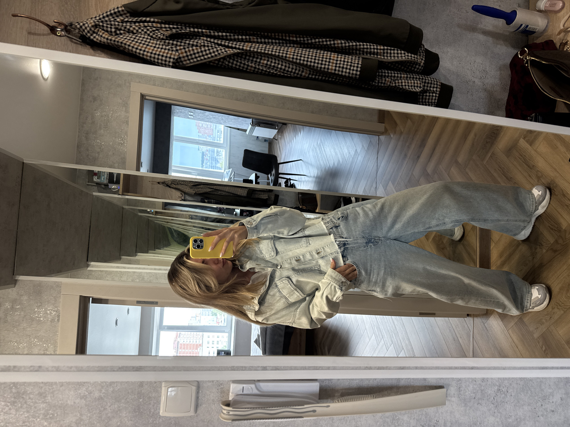 A person is captured taking a mirror selfie in an indoor setting, likely a home. The individual, with blonde hair, is wearing a light-wash denim jacket, light-wash wide-leg jeans, and white sneakers with grey accents. They are holding a yellow smartphone with a triple-lens camera setup, obscuring part of their face as they snap the photo.
The reflection in the mirror reveals various elements of the room. Above the person's reflection, a coat rack holds a dark jacket and a plaid brown and black checkered jacket. To the right of the coats, on a shelf, a blue lint roller, a small pink container, and a dark red or maroon bag are visible. Below the person's reflection, a grey countertop or shelf holds a white, ridged object, possibly a utility holder.
Through an open doorway in the background, a room with modern light-colored wooden flooring, laid in a herringbone pattern, is visible. A sleek black chair is positioned near a large window, which offers a view of a cityscape under a bright, clear sky, indicating it is daytime. The walls surrounding the mirror appear to be a light grey with some texture, and an oval-shaped wall light fixture provides additional illumination. The overall ambiance suggests a modern and well-lit interior. There is no discernible text visible in the image.