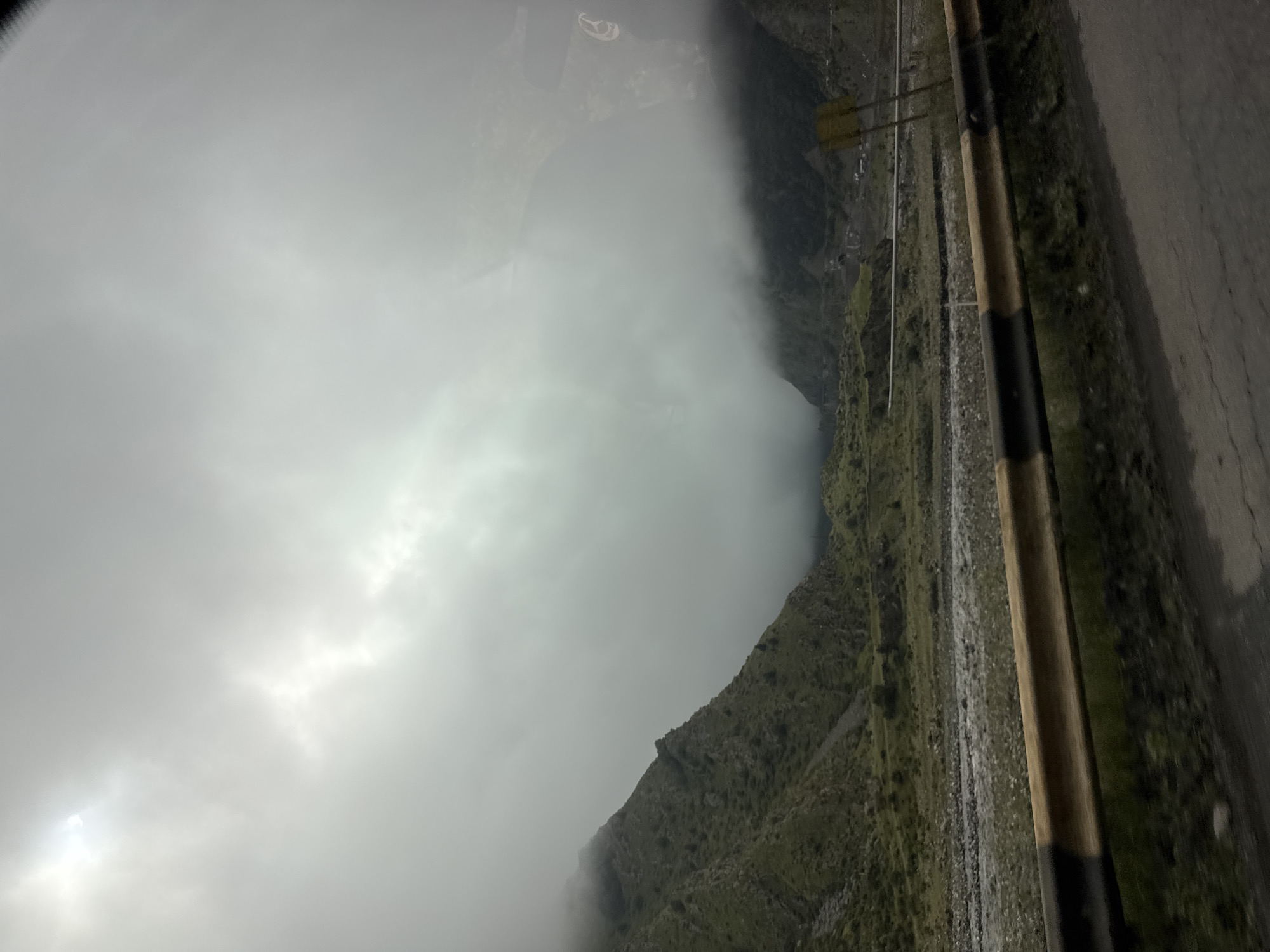 The image depicts a mountainous landscape with a river in the foreground. A yellow and black guardrail runs along the side of the road. The sky is overcast with clouds, and the sun is partially visible through the clouds. The mountains are covered in green vegetation. The scene appears to be captured from a moving vehicle, suggested by the motion blur in the foreground. The location is the city of Tsdo, Georgia, a mountainous region known for its scenic beauty. No people are visible in the image.