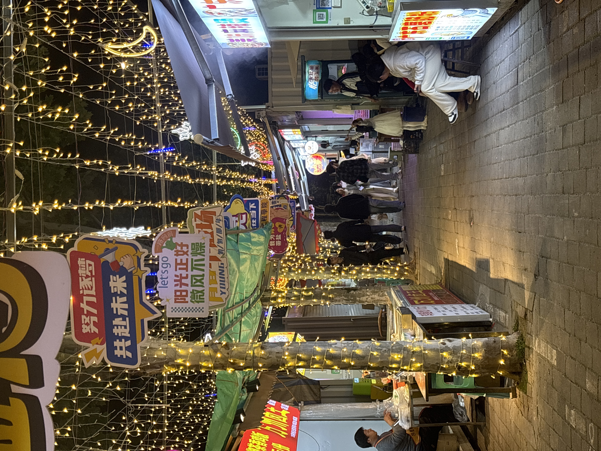 The image shows a narrow street at night in Jiangjin, China, lined with shops and adorned with strings of small, warm-toned lights. Pedestrians are walking through the street, some visible in the distance and others closer to the camera. Various signs are visible, some in Chinese characters, advertising the shops. One sign reads "努力逐梦 共赴未来" which translates to "Strive for dreams and jointly create the future." Another says "阳光正好 微风不燥." Which translates to "The sun is just right and the breeze is not dry." A person is seated near the lower part of the image, appearing to be attending to a stall. The street is paved with grey bricks. The atmosphere is festive and lively.