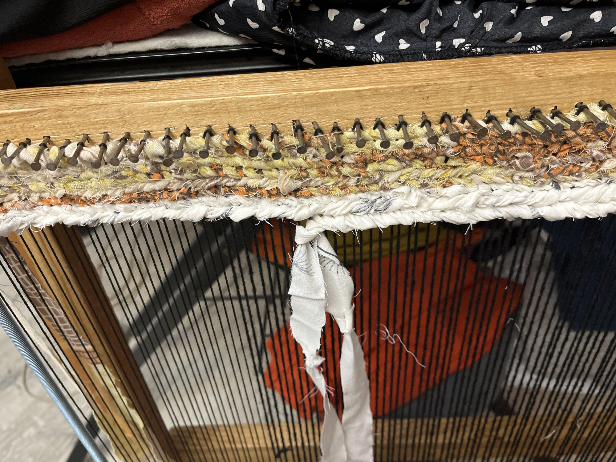 The image shows a close-up of a homemade weaving loom, likely in a domestic setting. The loom is constructed from wood, with nails hammered into the top edge to create a tensioning system for the warp threads. The warp threads are thin and black, creating a visible grid. The weft material is made of strips of fabric in various colors, including white, yellow, orange, and brown, some of which appear to be braided or twisted. A piece of white fabric is tied around some of the warp threads in the foreground. In the background, there is a glimpse of clothing with a black and white heart pattern. There are no people visible in the image. The scene does not provide any information that would allow for its localization to Krasnyye Barrikady, Russia, or any other specific city. The image appears to be taken indoors, possibly in a workshop or craft room.