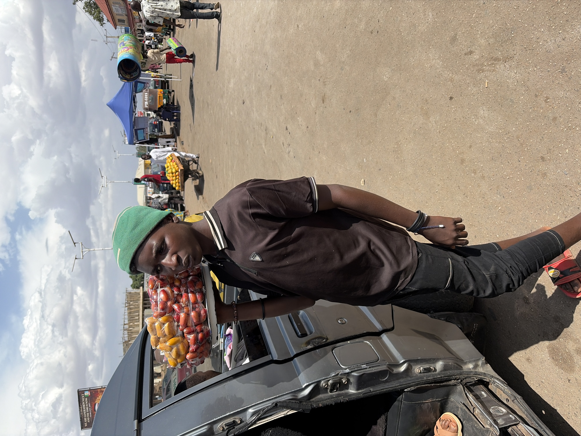 The image captures a vibrant street scene in Jos, Nigeria, on a bright, partly cloudy day. The ground is a light brown, possibly paved, and appears dusty.
In the foreground, a young Black man, the primary subject, is depicted from the waist up, leaning towards the camera while interacting with a vehicle. He wears a dark brown short-sleeved polo shirt with white trim, black cropped pants, and red sandals. A green knit beanie covers his head, and he has several dark bracelets on his left wrist. He holds a white tray filled with small plastic bags, each containing fresh fruit, appearing to be dates (both reddish-brown and yellow varieties). He is positioned next to an open grey vehicle, likely a car or a small public transport vehicle, with a person's foot in a light brown sandal visible inside.
The background reveals a bustling urban environment. Several other people are present, some walking, others standing or tending to stalls. Notable elements include:
- A blue canopy tent in the mid-ground.
- A person pushing a handcart laden with yellow fruits, possibly citrus.
- Another vendor with a large, colorful cylindrical object.
- Utility poles and overhead wires crisscrossing the sky.
- Buildings with a red-tiled roof and visible construction scaffolding in the distance.
The scene suggests active street vending and market activity. Shadows on the ground indicate a sunny day, likely mid-morning or early afternoon.
Visible text includes:
- A sign or banner reading "WE ARE GENEROUS NEXT AND GENEROUS IS NOW," with a date "15 JUNE 2022" partially legible below it.
- "PD.6082" visible on a box or object near the blue tent.
The overall impression is one of daily commerce and community life in a Nigerian city.