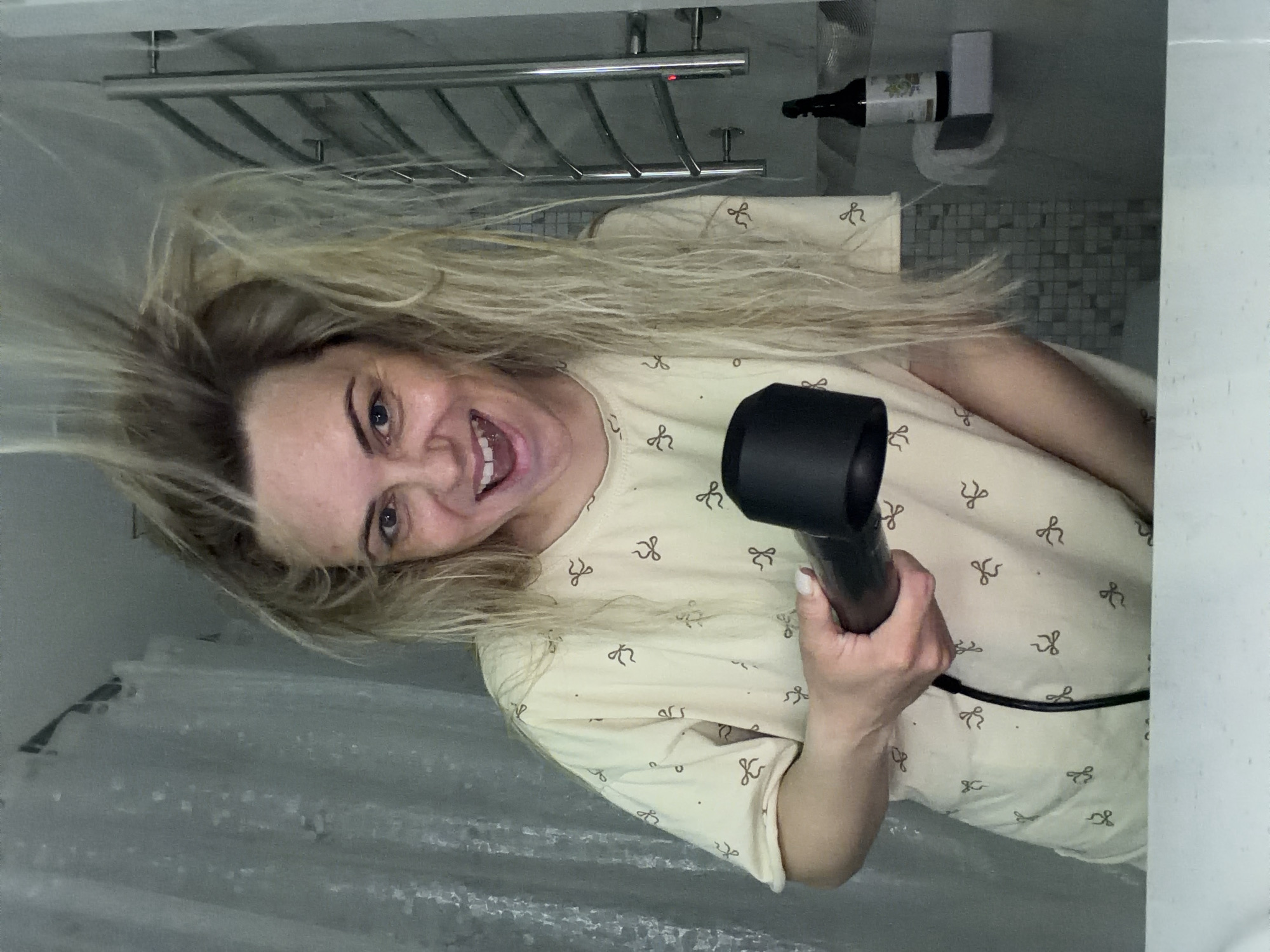 A woman is in what appears to be a bathroom, holding a black hair dryer pointed towards her face. Her blonde hair is disheveled, and she has a wide smile, showing her teeth. She is wearing a light yellow t-shirt with a pattern of small bows. A shower curtain is partially visible in the foreground. In the background, a chrome towel rack is mounted on the wall above her, along with a bottle and some other fixtures. There's no evidence to confirm or deny the location in Votkinsk, Russia.
