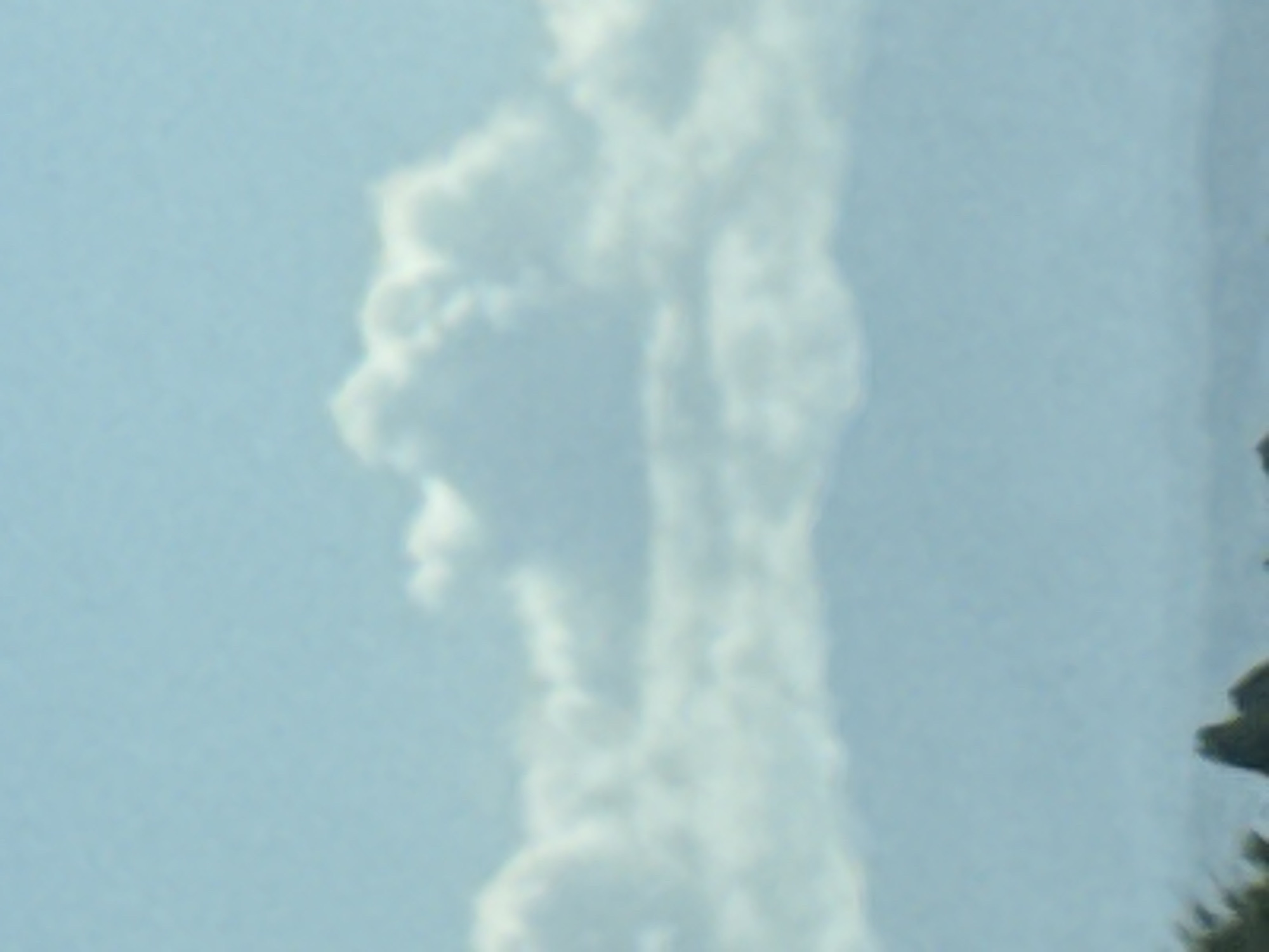A slightly blurred, medium-range shot captures a pale blue sky with scattered white clouds. Below the clouds, a hazy, dark mountain range stretches across the horizon. The lower portion of the image shows a dark, indistinct foreground with what appear to be trees. The scene suggests an outdoor environment, possibly in a mountainous region like Chim-Korgon, Kyrgyzstan, with clear but overcast weather conditions.