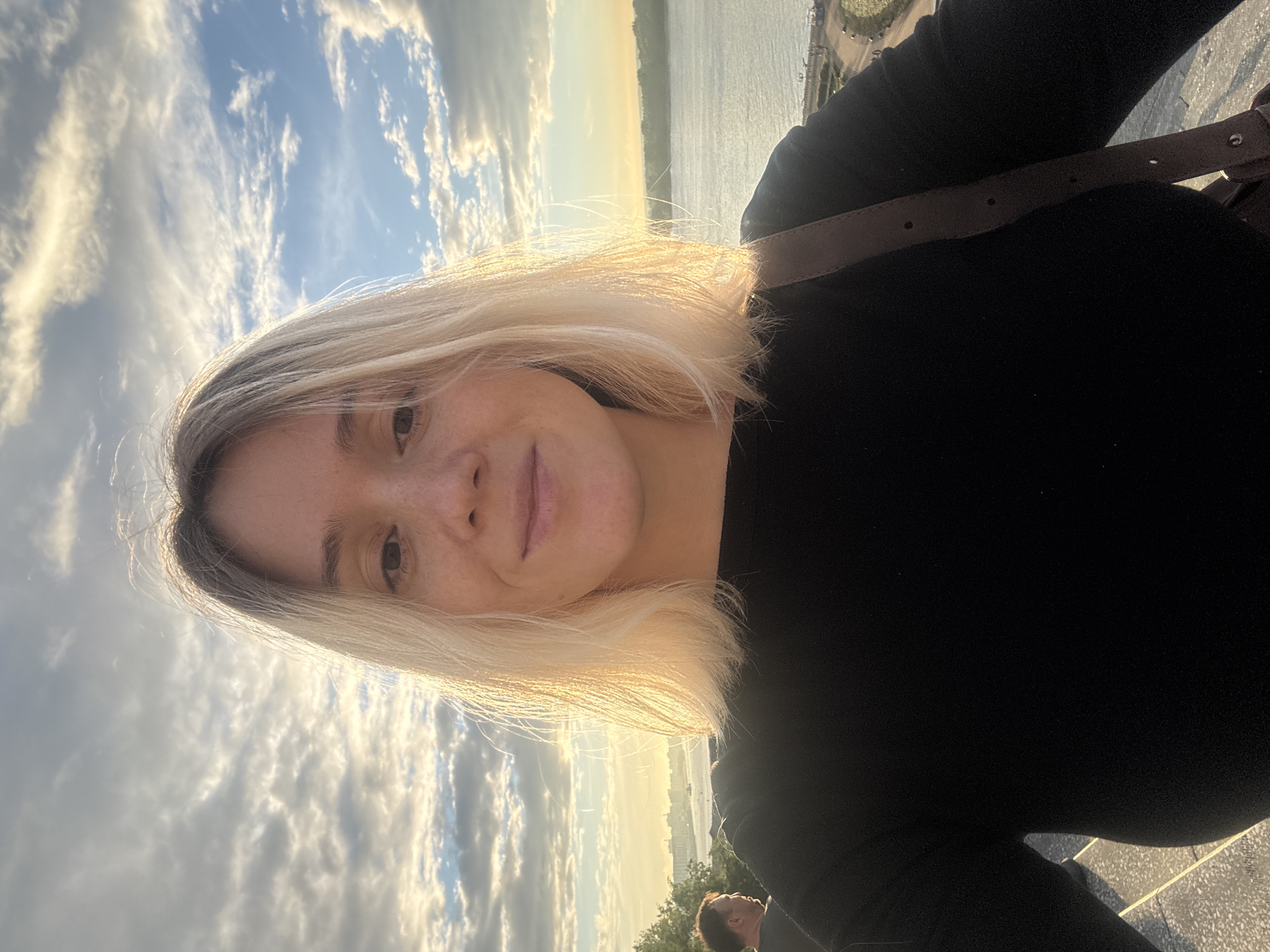 A medium-shot selfie is taken by a young woman with blonde hair, freckles, and a slight smile, looking directly at the camera. She is wearing a black long-sleeved shirt. Behind her, a body of water is visible, likely the Volga River, with a cityscape and green treeline in the distance. The sky is filled with dramatic clouds, illuminated by the golden light of either sunrise or sunset. In the background, to the left, a man is seen from the side, also outdoors. The scene suggests an outdoor location in Nizhny Novgorod, Russia, possibly a viewpoint overlooking the river.