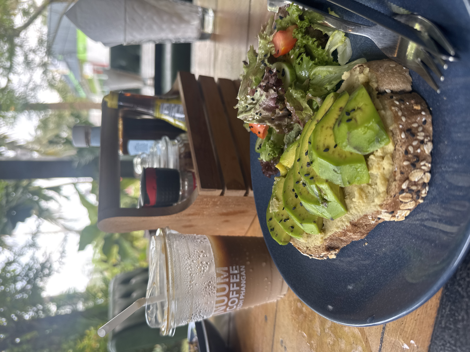 On a wooden table in Baan Kai, Thailand, a partially eaten plate of avocado toast with a side salad sits next to a glass of iced coffee. The toast is topped with sliced avocado and black sesame seeds, and the salad contains lettuce and chopped tomatoes. A fork is placed on the plate, ready to eat. A wooden stand holds various bottles and containers. The foreground features a cup of coffee with condensation and a plastic straw. The background is blurred, showing greenery and structures, indicating an outdoor setting, likely a cafe or restaurant.