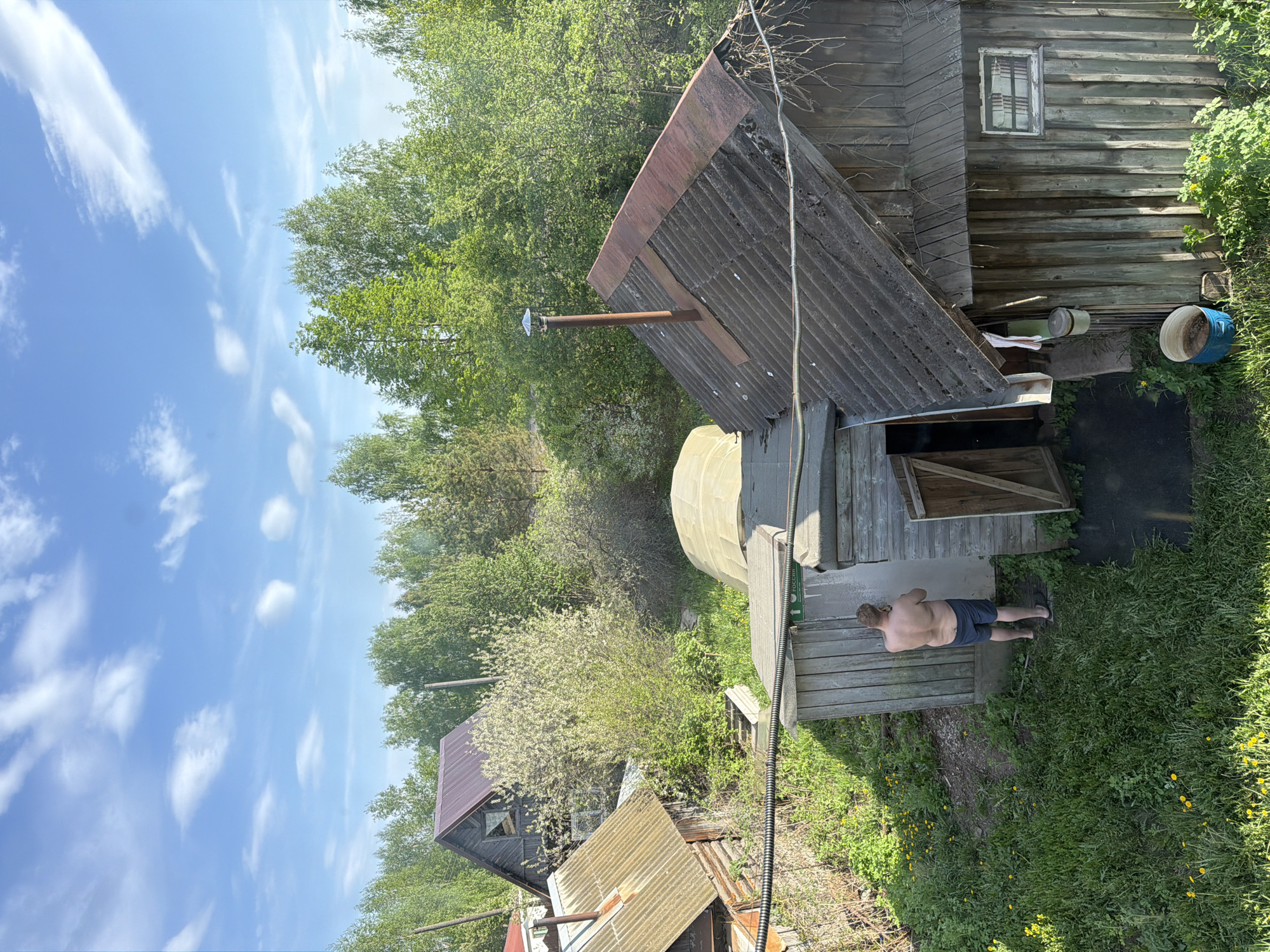 A medium-angle daytime shot depicts a rural setting possibly within or near the city of Plamya, Russia, based on visible signage in the Cyrillic alphabet. The image is slightly tilted counter-clockwise. A shirtless man in dark shorts and sandals stands on a patch of grass outside a weathered wooden structure, possibly a sauna or bathhouse, with an open door. The structure has a corrugated roof with a metal chimney. Adjacent to this is a metal tank or reservoir. Further back and slightly below, another aged wooden building is partially visible. The surrounding area is overgrown with grass, weeds, and dandelions. Trees are visible in the background. The sky is blue with scattered clouds. A power line runs across the lower portion of the frame.
