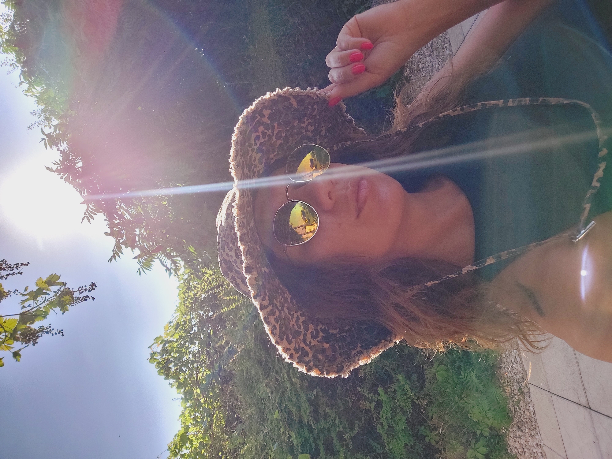A woman with long, wavy brown hair is wearing a leopard print bucket hat and round sunglasses. She is holding the brim of her hat with her right hand, which has bright pink fingernails. She is looking at the camera with her mouth slightly open. The background is a lush green garden with dappled sunlight. The scene appears to be outdoors, possibly in the city of Sozopol, Bulgaria, given the Mediterranean vegetation. The overall impression is one of a relaxed, sunny day.