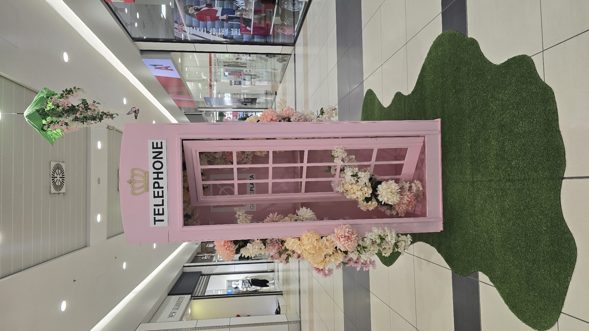 An indoor scene within a mall corridor at Mall Galleria Burgas, Bulgaria, features a decorative, light pink British-style telephone booth as its central subject. The booth is positioned on an irregularly shaped piece of green artificial turf on the tiled floor. Artificial flowers in shades of pink, white, and pale yellow profusely adorn the exterior and interior of the booth; its door is slightly ajar, revealing more floral arrangements inside. A gold-colored crown emblem is mounted above the word "TELEPHONE" in black capital letters on the booth's top section. Overhead, an inverted green umbrella, suspended from the white paneled ceiling, drapes artificial pink and white flowers and green foliage. Two artificial butterflies, one purple and one orange, are suspended nearby. The ceiling is equipped with recessed lighting and a circular air vent. Storefronts line both sides of the corridor: "NEW SILHOUETTE" with mannequins is visible on the left, and on the right, stores display items behind glass, including one with an "A1" logo and another featuring "ENZO NORI" branding and a large advertisement. The area is brightly lit. No individuals are clearly identifiable in the immediate foreground or midground.