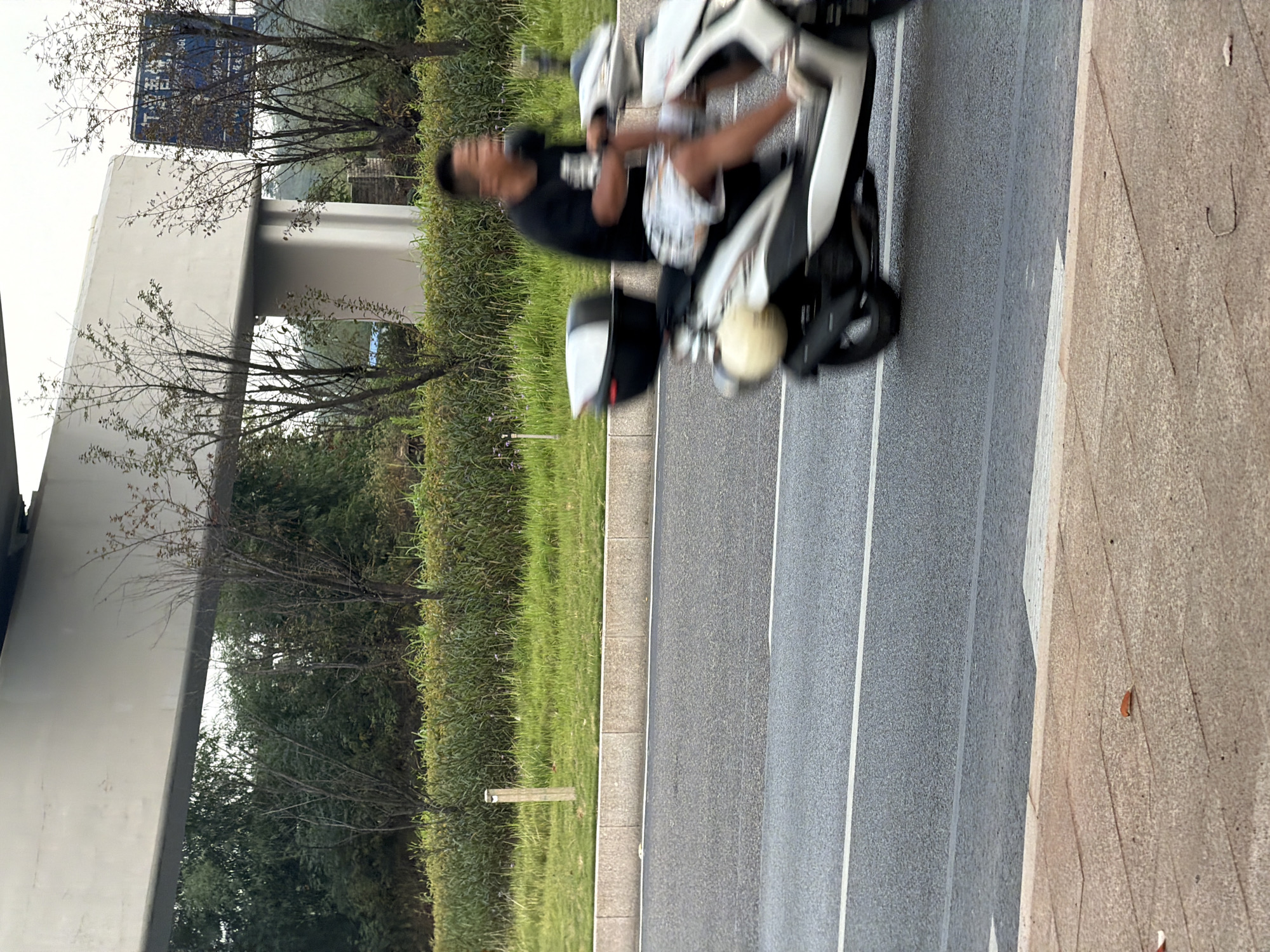 A young man wearing a black t-shirt and patterned shorts rides a white and black scooter on a paved road. He is in motion, with his face blurred and mouth open. The scooter has a black storage box on the back. The road has white lane markings. In the background, there is a green grassy area with some bushes and trees. An elevated concrete structure spans across the scene above the trees. A blue sign with Chinese characters is visible on the right side, attached to the elevated structure. The foreground shows a textured paved walkway. The overall scene appears to be in an outdoor urban environment, likely in the city of Jiangjin, China, judging by the presence of Chinese text on the sign.