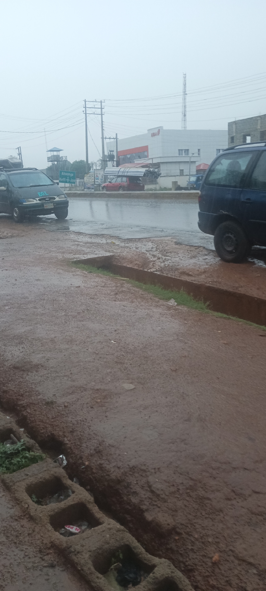The image shows a street scene in Akwanga, Nigeria on a rainy day. The road is wet, and puddles are visible. In the background, there is a building with "UBA" written on it. Street signs are also visible, one indicating directions to "Akwanga, Keffi, Jos". Several vehicles are present, including a black car with items on its roof. The foreground shows a muddy, unpaved area with concrete drainage blocks, some filled with debris. A communication tower is seen in the distance. There are no people visible in the frame.