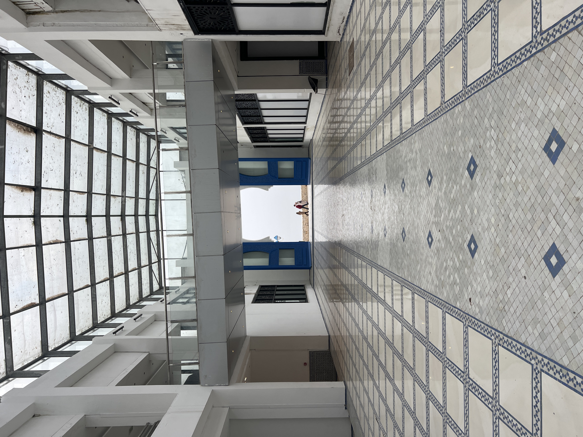 The image displays a long, brightly lit corridor or covered walkway, characterized by a blend of modern and traditional architectural elements. The perspective looks down the length of the hallway towards an open outdoor area.
On the left, a large glass-paneled ceiling or skylight extends overhead, allowing natural light into the space. The glass appears somewhat aged or dirty. Below this, white beams and sturdy grey/silver pillars form part of the building's structure.
The floor is extensively tiled with a striking white and blue geometric pattern, creating a sense of depth and design. The right wall is also adorned with large white tiles bordered by intricate blue geometric motifs, echoing the floor's aesthetic. Further along the right side, black decorative grates or panels are visible high on the wall or ceiling, possibly serving as ventilation or design elements.
At the far end of the corridor, a prominent, vibrant blue archway or structure provides an exit, leading to a bright outdoor area. Two small, indistinct figures are visible in the distance beyond this archway, with one appearing to wear red clothing. A small, light-colored object, possibly a balloon, is also seen floating or positioned in the outdoor space.
The scene is bathed in daylight, indicating a daytime setting with clear weather outside. The architectural style, particularly the blue accents and geometric tiling, suggests a Moroccan influence, consistent with the given location context of M'Diq. The corridor itself is devoid of activity, presenting a calm and spacious environment. No discernible text is visible in the image.
