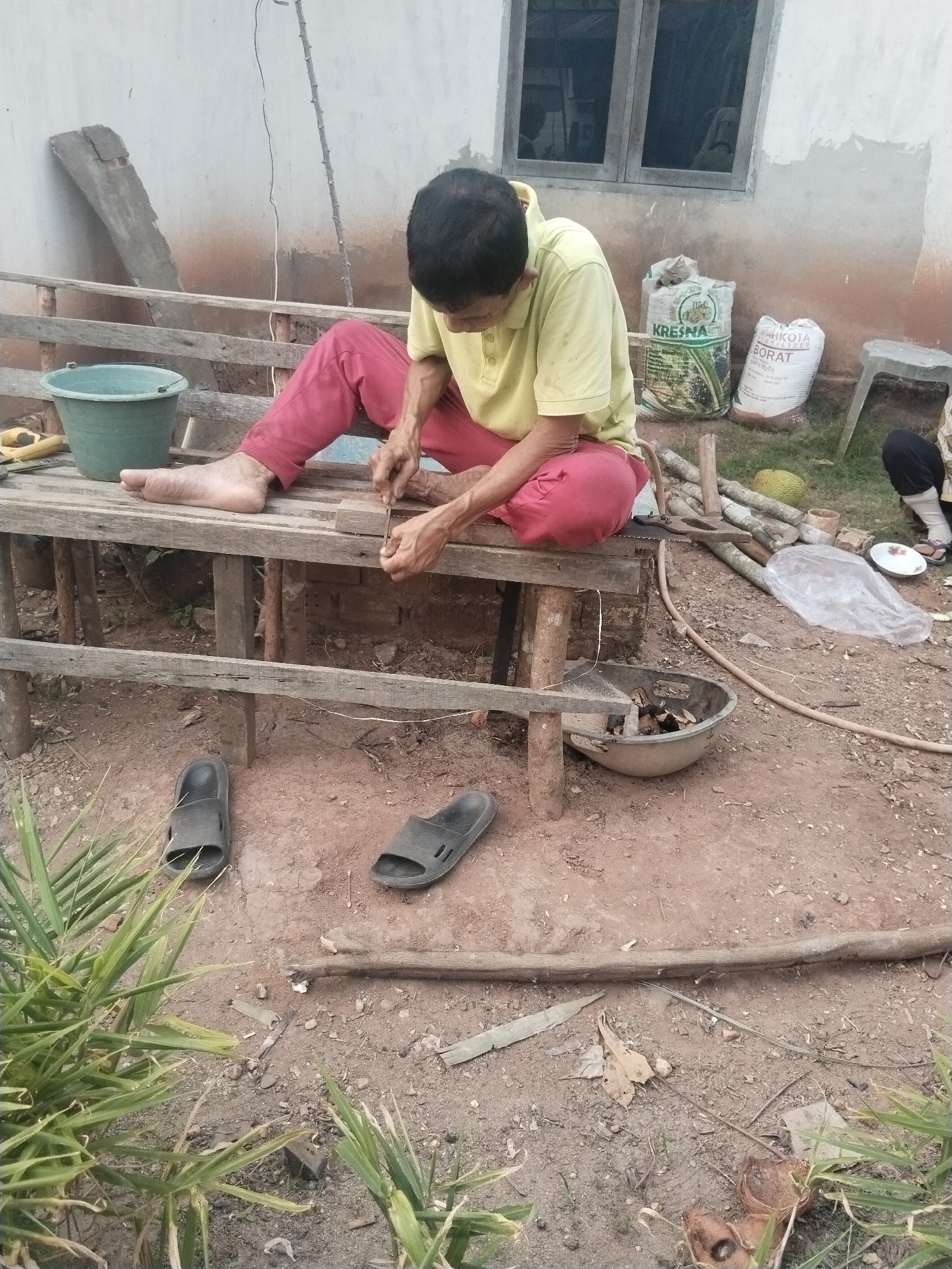A person wearing a yellow polo shirt and red pants sits on a wooden bench, engaged in a task that appears to involve carving or smoothing a small wooden block with a tool. The individual's focus is directed downwards towards their work. To the left of the person, a green plastic bucket rests on the bench. In the background, a building with a window and a rough concrete wall is visible. Several sacks, including ones labeled "KRESNA" and "MAHKOTA BORAT," are placed near the building. The ground is covered in dirt and scattered with debris, including pieces of wood and plant matter. A pair of black sandals are on the ground in front of the bench, and another pair is visible further to the right. A durian fruit and a plastic bag are also present in the scene. The environment suggests an outdoor setting in a rural or semi-rural area. Based on the presence of the sacks with Indonesian labeling and the overall setting, the location is likely Suka Ramai, Indonesia.