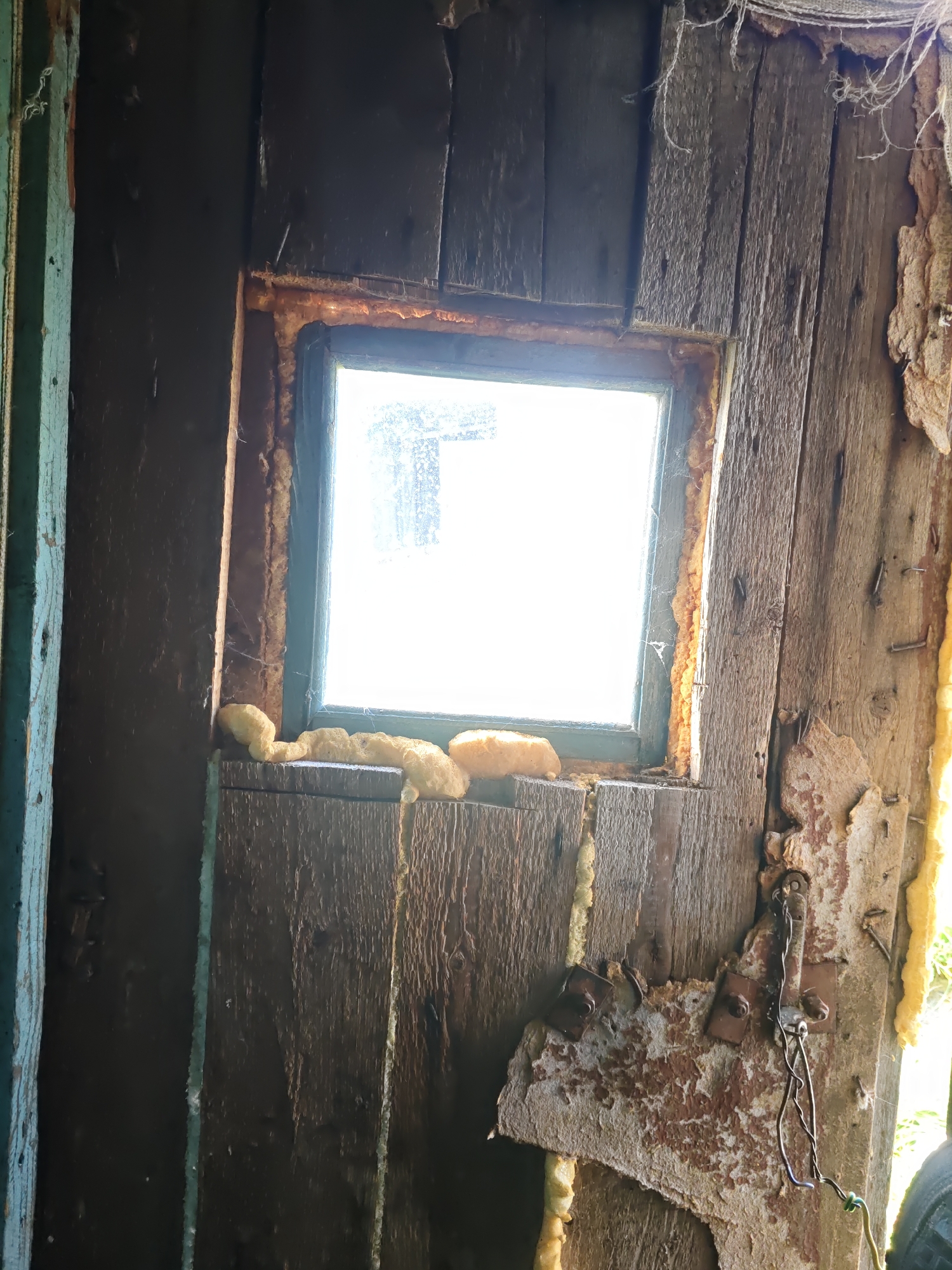 This image depicts the interior of a dilapidated wooden structure. A small, grimy window with a bright, blown-out background is centrally located, surrounded by expanding foam insulation that has been applied unevenly. The walls are constructed of rough, weathered wooden planks, with evidence of peeling paint and decay. To the right of the window, a piece of what appears to be a metal latch or hinge is attached to the wooden wall, secured by rusty screws. Wire is also attached to this metal piece. The overall impression is one of disrepair and neglect within the structure. The location is identified as Платошино, Russia, implying this structure is situated within that area.