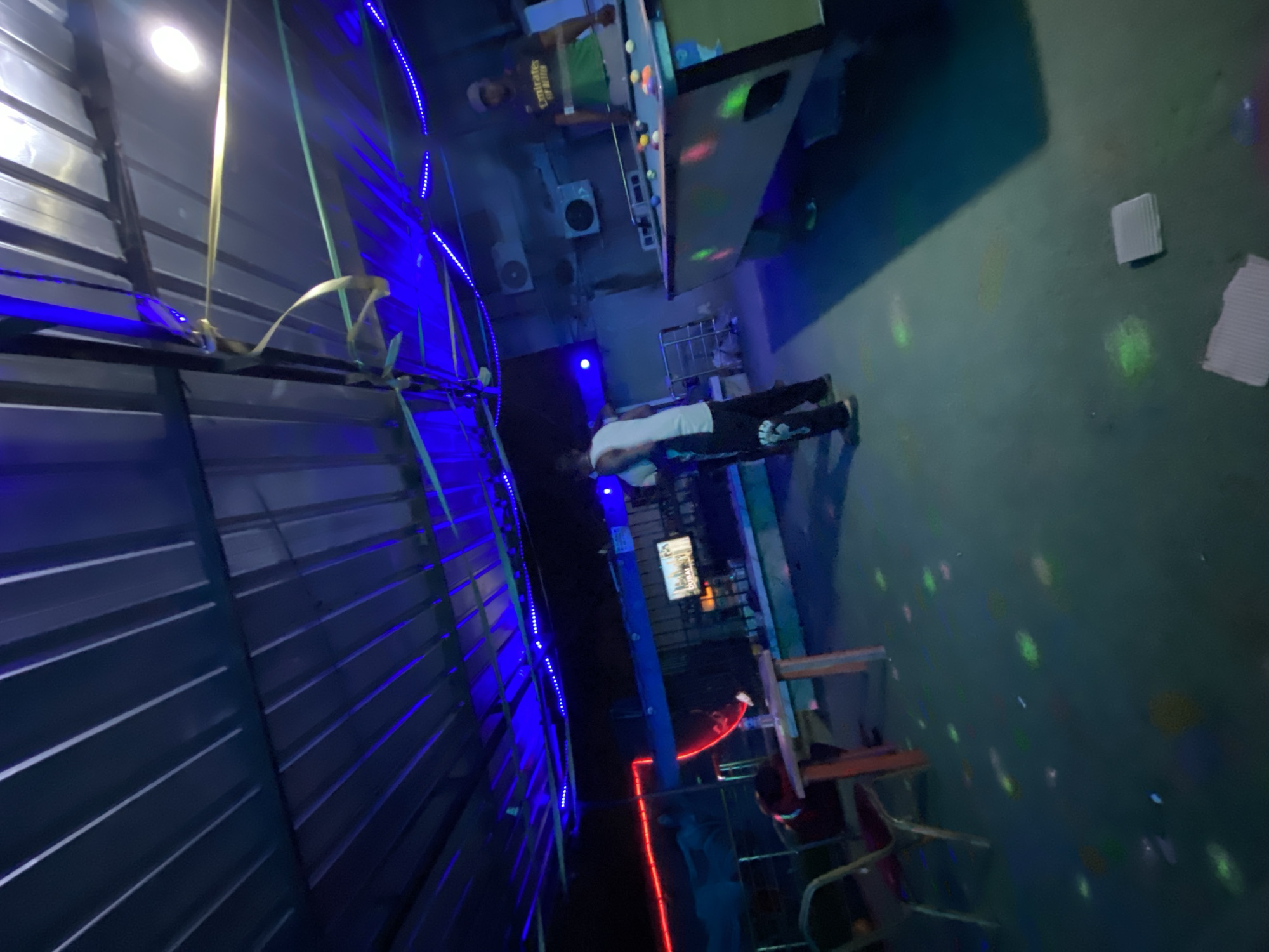 The image shows an indoor scene in Ado, Nigeria. There are two individuals visible. One is playing pool at a pool table, while the other is standing nearby. There are tables and chairs, and what appears to be a corrugated metal structure with blue neon lights. There is also a wall with disco-style lights.