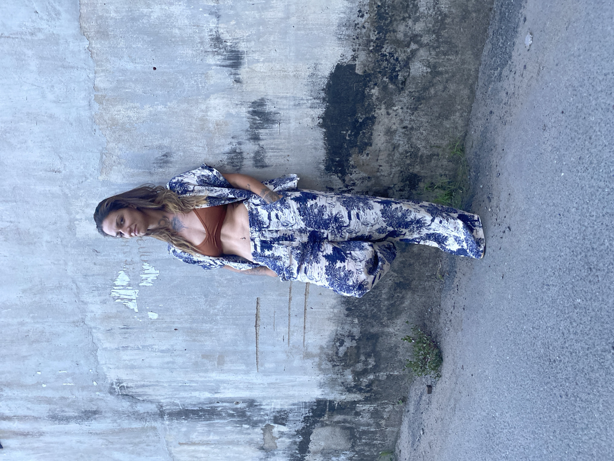A woman stands in an alley, leaning against a concrete wall in Đồng Nai, Vietnam. She wears a brown top and a blue and white patterned pajama set. The wall has peeling paint and dark stains. A paved surface is adjacent to the wall, showing minor plant growth.