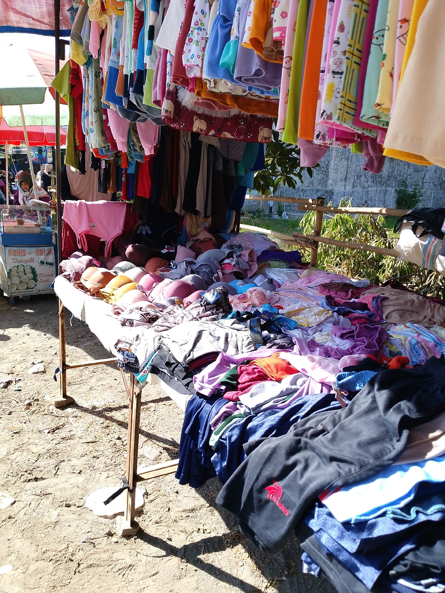 This image depicts an outdoor market stall in Tegalwangi, Indonesia. The stall displays a variety of clothing items, primarily undergarments and children's clothing, for sale. In the foreground, a table is laden with bras in various colors and styles, along with folded piles of fabric. To the left, a woman wearing a headscarf is visible, seemingly attending to the stall. Behind the table, numerous colorful children's garments hang from a rack, exhibiting cartoon prints and bright hues. The ground surrounding the stall is unpaved and dusty, with some debris scattered about. The market appears to be set up in a sunny outdoor location, with a wall and vegetation visible in the background.