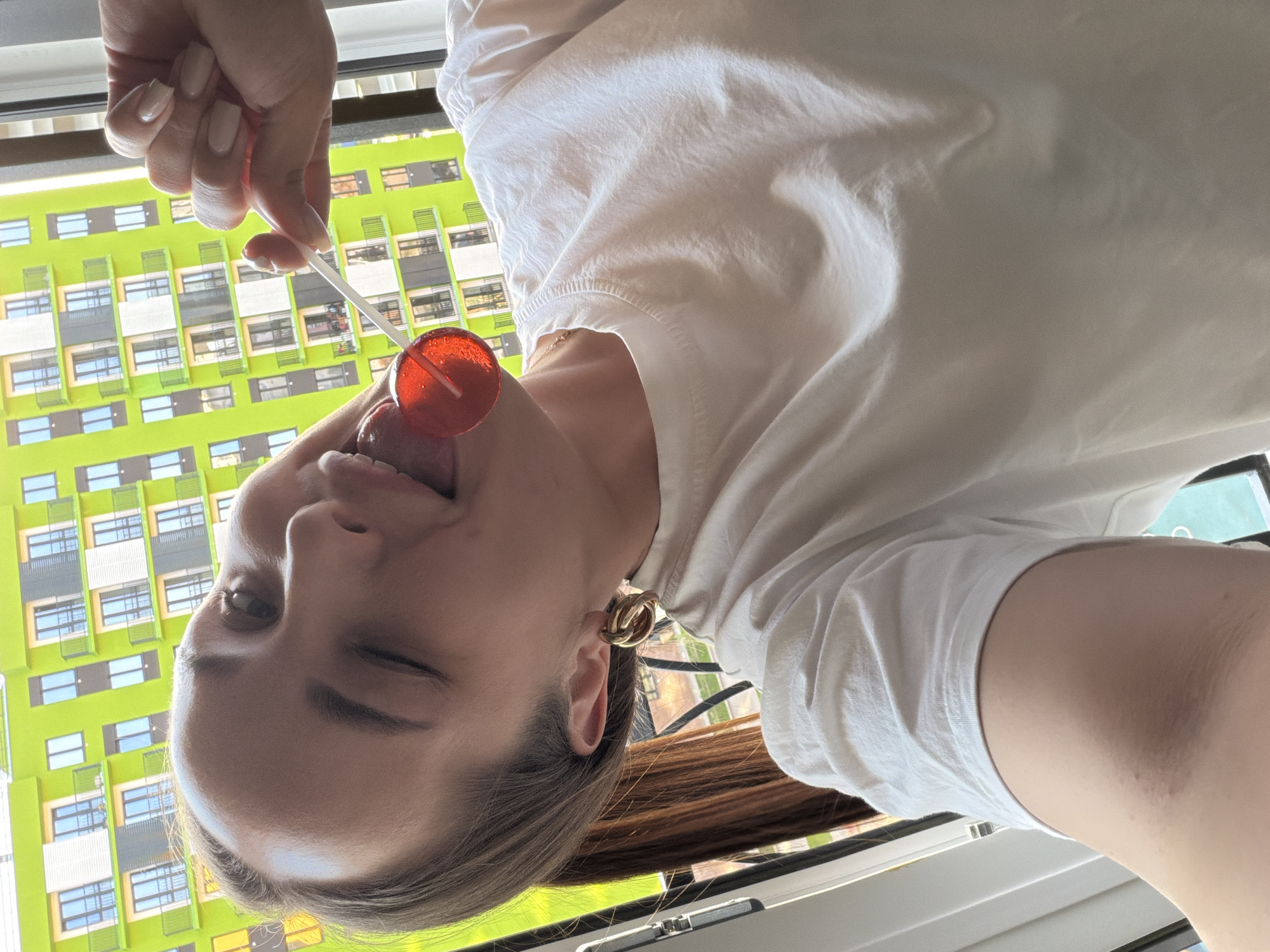 A young woman holds a red lollipop to her mouth, her tongue slightly extended. She wears a white shirt and a gold earring. A building with a green facade and numerous windows is visible in the background. The location is Pervomaysky, Russia.