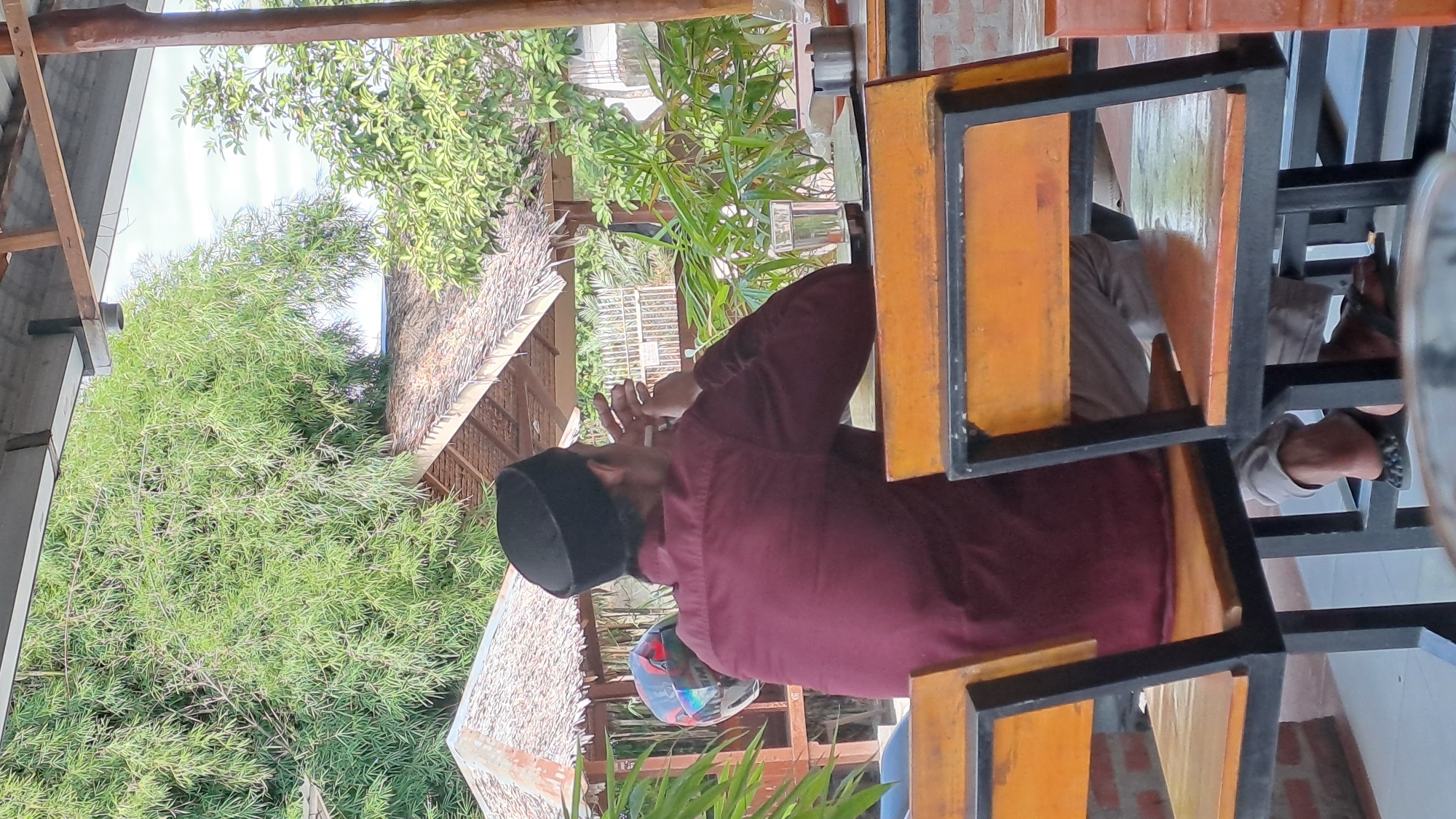A man wearing a maroon long-sleeved shirt and a black peci hat sits at a table outside under a wooden overhang. He is facing away from the camera and appears to be smoking a cigarette, holding it in his right hand near his face. He is wearing light-colored pants and dark sandals. In front of him, on the table, are a glass of water, a notebook, and an ashtray. Behind the man, a motorcycle helmet is visible. The background consists of lush green foliage, including bamboo, and thatched-roof structures. The overall setting suggests an outdoor seating area, possibly a cafe or restaurant, in Julikeude Dua, Indonesia.