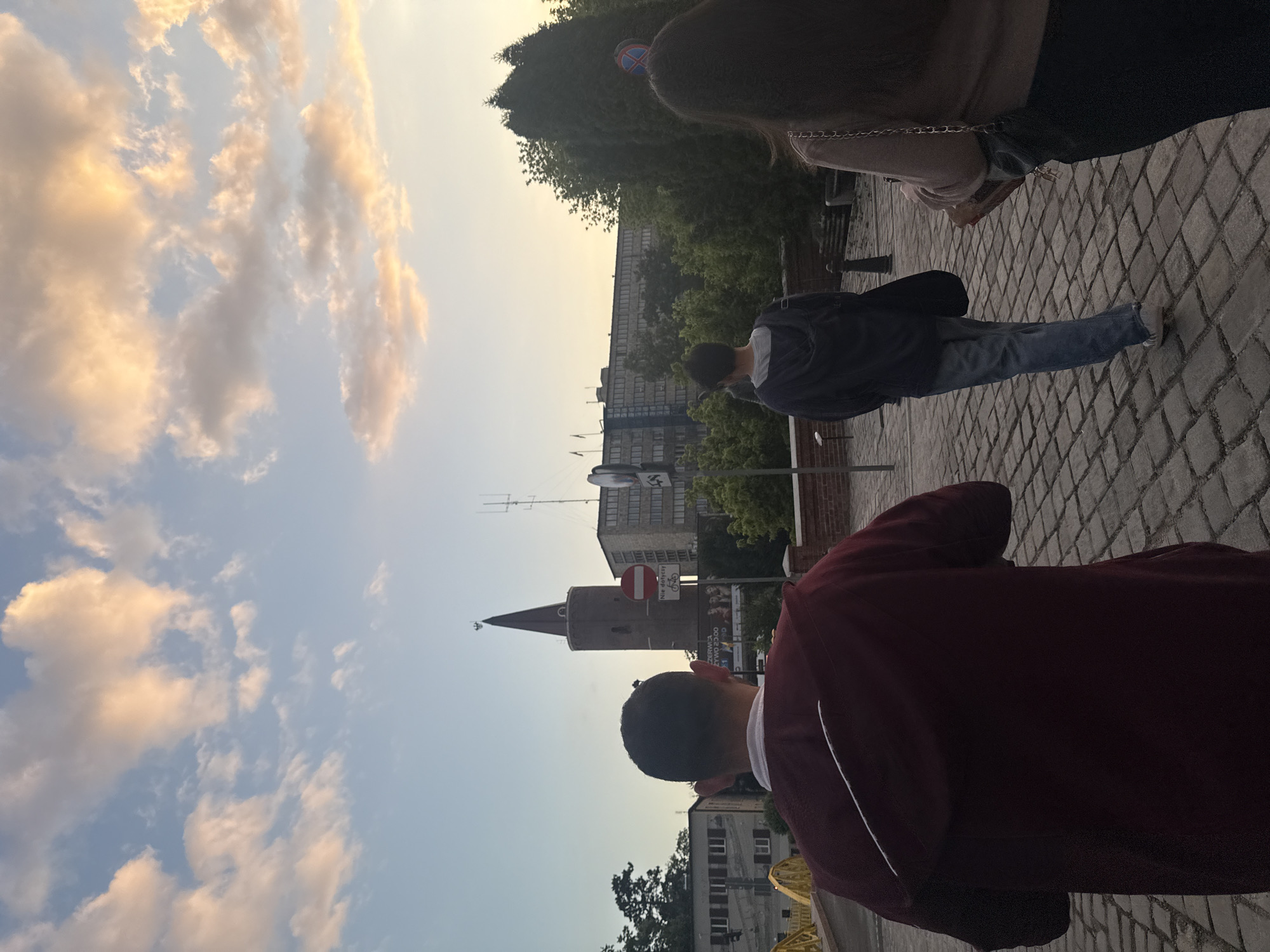 This eye-level outdoor photograph captures a street scene in Opole, Poland, during the golden hour. The expansive sky occupies the upper portion of the frame, showcasing soft, warm-toned clouds illuminated by the late afternoon or early evening sun. A small airplane is faintly visible in the distant sky.
In the foreground, the back of a person wearing a dark maroon jacket with a white stripe is visible, standing on grey cobblestone pavement. Slightly further back, another individual in a dark blue jacket and blue jeans walks along the same pavement, also seen from behind. A third person is partially obscured in the midground.
The background prominently features the historic brick Piast Tower with its conical roof, a recognizable landmark of Opole. Surrounding the tower are modern buildings, some topped with antennas, and lush green trees. Several traffic signs are visible near the tower; one red circular sign indicates "No Entry" (B-2) and another signifies "No Cycling" (B-9). On one of these signs, the Polish phrase "Nie dotyczy" (Does not apply to) is legible. Below, a billboard displays partially visible text, including fragments like "CZERW" and "ESTUDIO". A yellow metal structure, possibly a bridge railing, is seen in the bottom left. The overall lighting suggests a serene atmosphere as day transitions to evening.