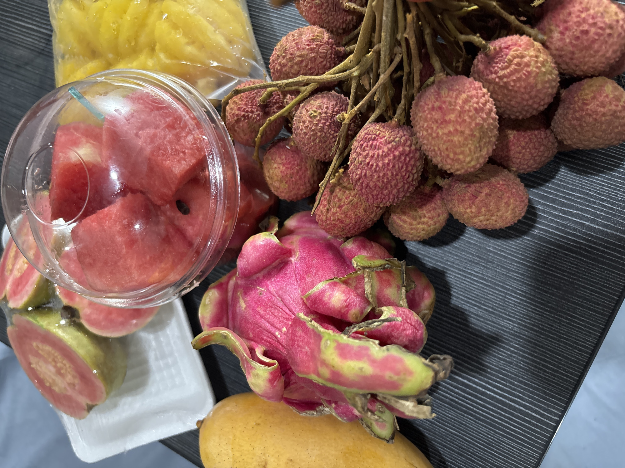 The image displays a variety of fruits arranged on a dark, horizontally lined surface. The fruits include: lychees still attached to their branches, cut pieces of watermelon in a plastic container, starfruit in a plastic bag, a dragon fruit, a mango, and a guava cut in half. The arrangement of the fruits, along with the specific fruits included, suggests the location is in a tropical climate, which is congruent with the location of บ้านบางรักษ์, Thailand.