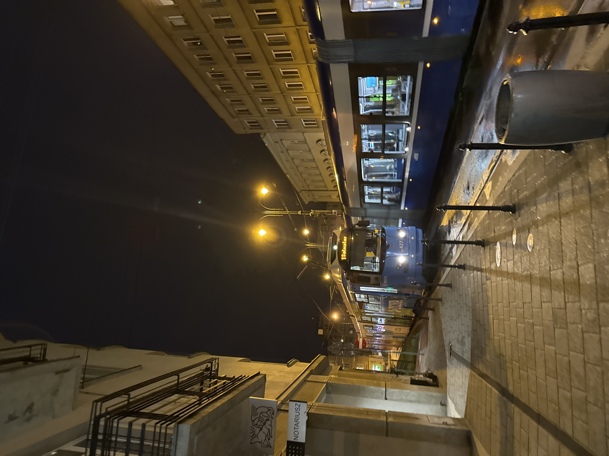 The image depicts a night scene in Krakow, Poland, likely along a tram line. A blue tram dominates the right side of the frame. The street is paved with brick, and there are street lamps and a trash receptacle. On the left side of the image, there is a building with a sign that reads "Notariusz," which translates to Notary. The darkness of the sky suggests it is late evening or night.