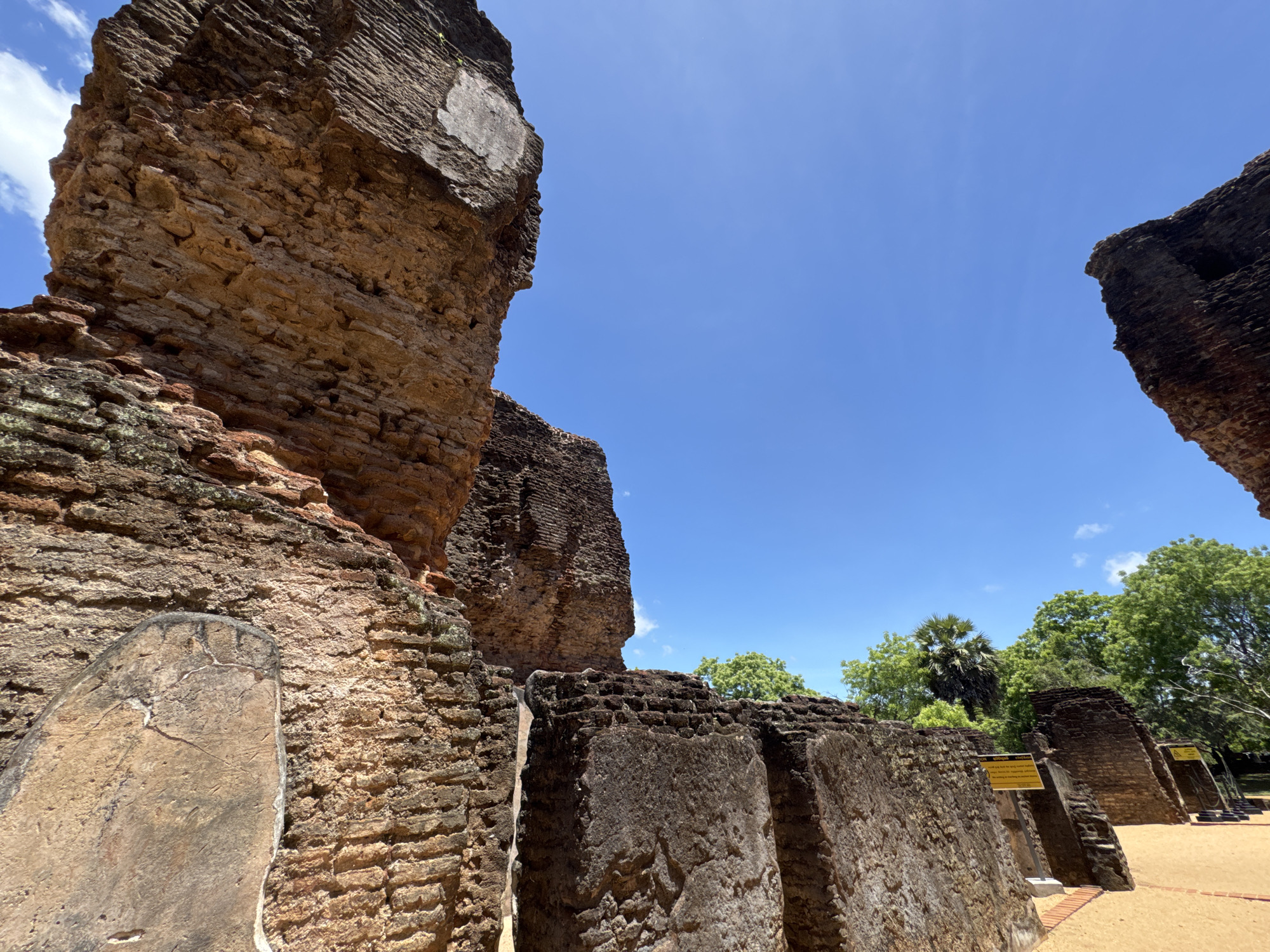 The image shows the ruins of an ancient structure in Polonnaruwa, Sri Lanka. The buildings are made of brick and stone, and have been weathered over time. There are trees and greenery in the background, and the sky is clear and blue. The scene is peaceful and serene.