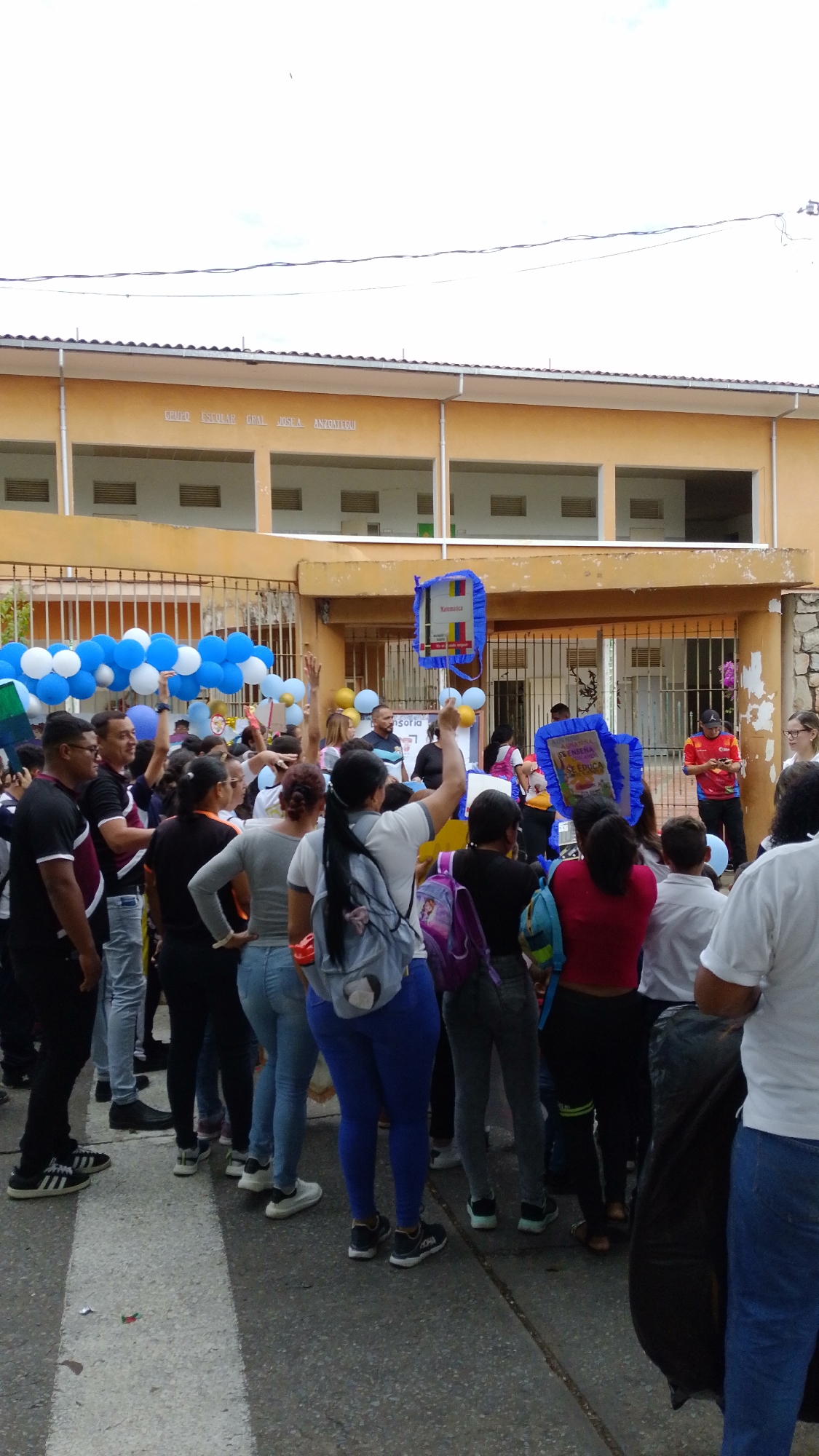 A daytime outdoor gathering is taking place in front of the Grupo Escolar Gpal. Jose A. Anzoategui school in Tinaquillo, Venezuela. A diverse crowd of people, including adults and likely students, fills the paved area. Many individuals are facing the school building, with some raising their hands in apparent engagement.
The event features decorations, including clusters of blue and white balloons on the left side of the scene. Several people are holding up large, blue, rectangular signs or themed props. One sign clearly displays the word "Matematica" (Mathematics), while another bears the text "LA SENDA SE EDUCA" (The Path is Educated/Learned).
The school building in the background is a light orange-yellow structure with white trim, featuring multiple open window or archways across its facade. A metal fence separates the crowd from the building. The sky is overcast, indicating a bright, cloudy day. A person in a red shirt stands near the building, possibly addressing or organizing the crowd. The overall scene suggests a lively school event, such as an assembly, celebration, or educational fair.