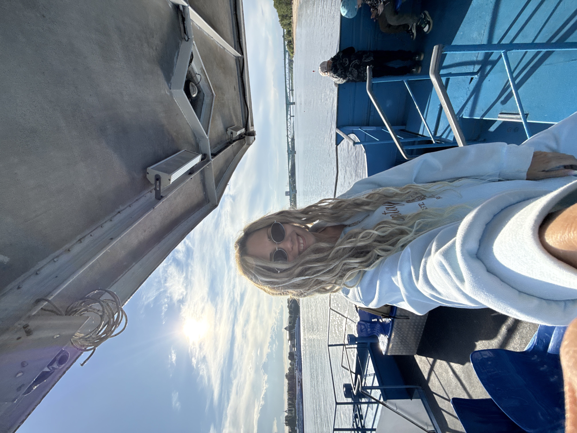 The image shows a woman with blonde dreadlocks and sunglasses taking a selfie on a boat. She is wearing a light blue hooded sweatshirt. The boat has blue railings and seating. In the background, there is a body of water, likely the Volga river, and a view of the city of Заволжье, Russia, with a bridge visible in the distance. There are other people on the boat, including one individual wearing a patterned shirt and a light blue hat. The weather is sunny, with a clear blue sky and a bright sun casting shadows on the boat.