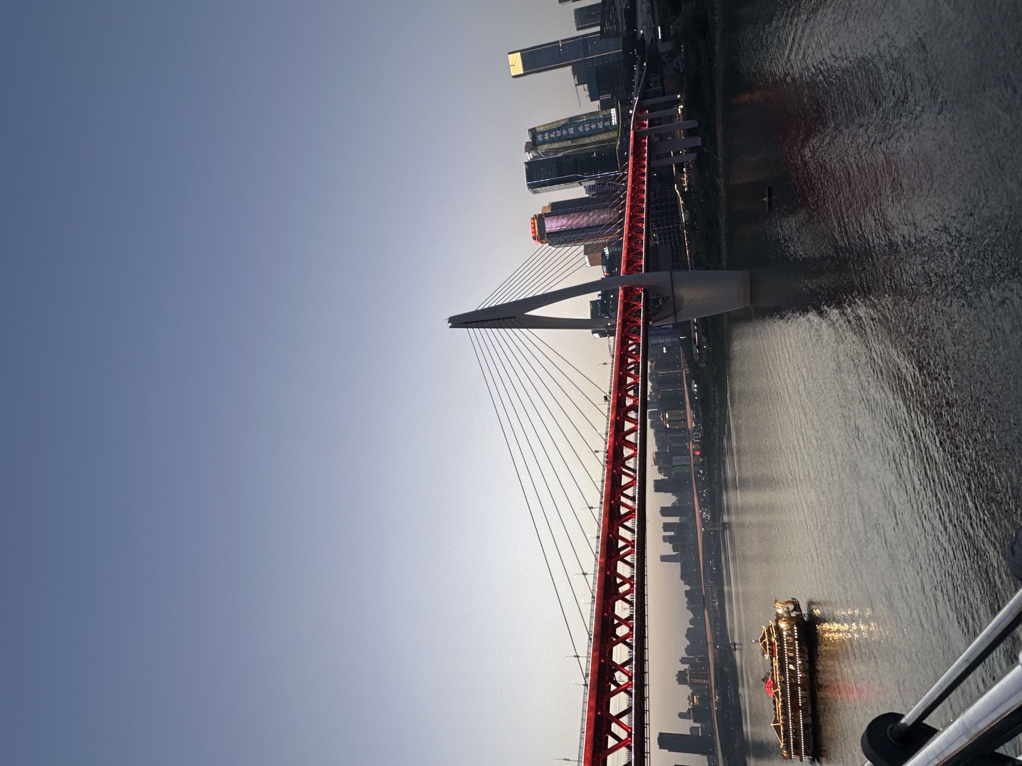This media file captures a twilight scene in Yuzhong District, China, showcasing a prominent red cable-stayed bridge spanning a wide river. The image is taken from a slightly elevated perspective, possibly from another bridge or high viewpoint, looking across the water towards a bustling cityscape.
The main subject is a striking red bridge with a tall, white, A-shaped pylon from which numerous cables descend to support the bridge deck. The bridge's truss structure is brightly illuminated in red, reflecting onto the water below. To the left, a portion of what appears to be a railing or structure from the viewer's location is visible.
On the far bank of the river, a dense cluster of modern skyscrapers forms a city skyline. Many of these buildings are lit up with various colors and patterns, typical of a vibrant urban nightscape. Visible text on one building facade includes "重庆国际金融中心" (Chongqing International Financial Center), and on another, "环球金融中心" (Global Financial Center), confirming the location as Chongqing.
The river itself is dark and expansive, with subtle ripples on its surface reflecting the city lights and the bridge's illumination. A large, illuminated passenger boat, adorned with strings of bright lights, travels slowly on the river, its glow creating streaks of light on the water.
The sky is a clear, gradient blue, transitioning from a lighter hue near the horizon, where the sun has just set or is about to rise, to a deeper blue overhead. The time of day is clearly dusk or early evening, characterized by the natural dimming light and the prevalence of artificial illuminations. The weather appears calm and clear, contributing to the serene yet impressive atmosphere of the urban landscape. There are no identifiable people visible, but the illuminated boat suggests ongoing activity.