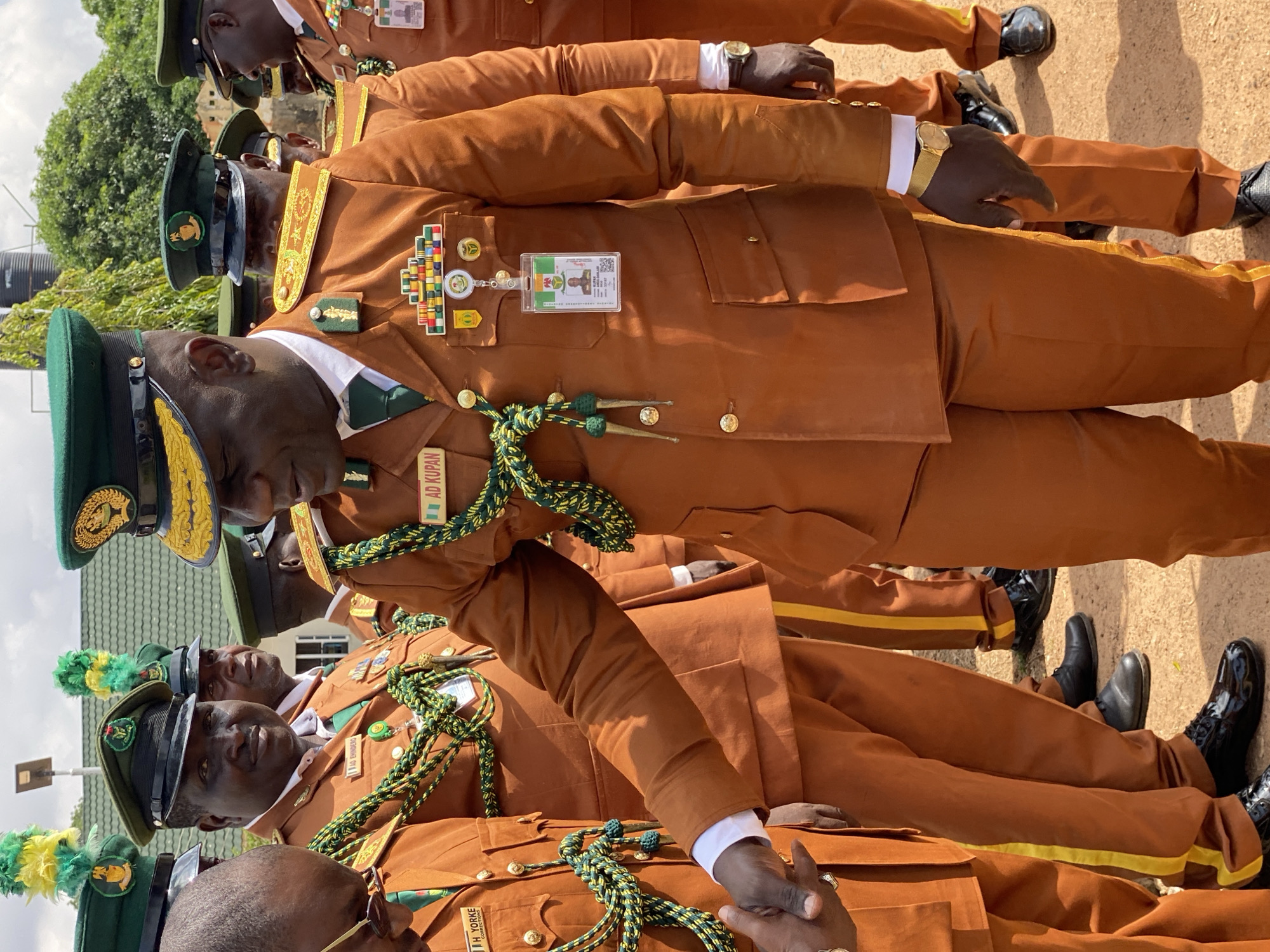 The image depicts a group of men in uniform, possibly correctional officers, standing outdoors. The uniforms are a distinctive burnt orange color with green and yellow accents. Several men are wearing peaked caps with green brims and ornate insignia. Some individuals have badges and medals pinned to their chests, and lanyards with identification cards are visible. One man in the foreground is shaking hands with another individual. The background shows trees and buildings, suggesting an outdoor setting in a populated area. The ground appears to be dirt or gravel. The overall atmosphere seems formal, possibly a ceremony or official gathering. The location is specified as the city of Kaduna, Nigeria.