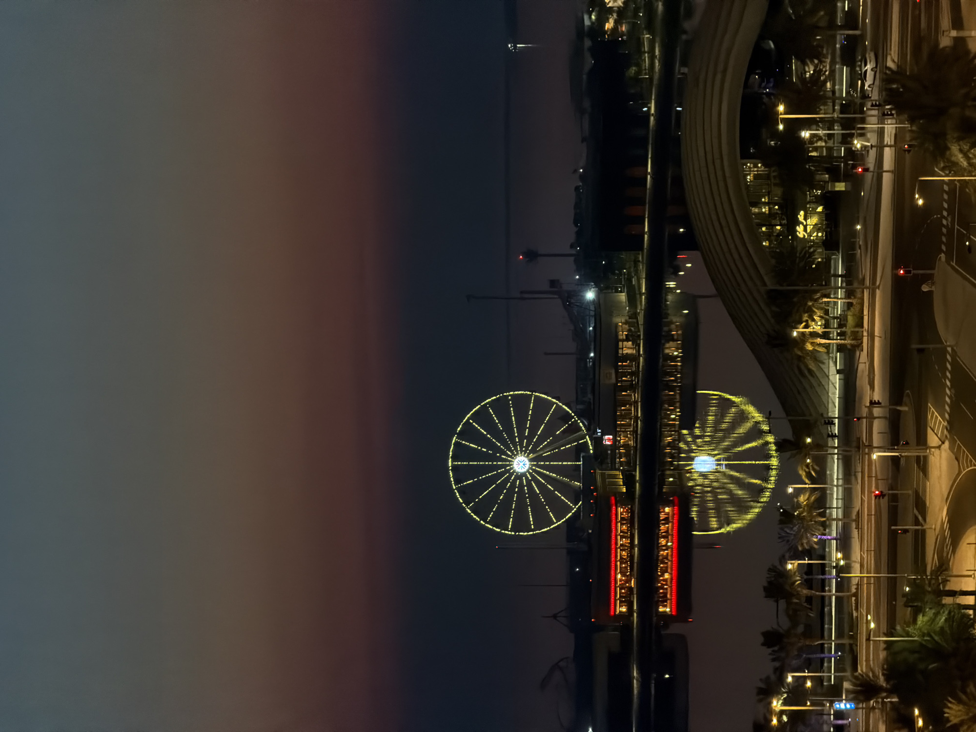 An aerial, high-angle, long shot captures a cityscape at dusk, with a Ferris wheel as the central illuminated subject. The Ferris wheel, adorned with yellow lights, dominates the mid-ground, its reflection shimmering in the dark water below. Adjacent to the Ferris wheel is a building with numerous red lights, also reflected in the water. The foreground features a street with traffic lights, crosswalks, and palm trees lining the sidewalks. The sky transitions from a soft gradient of twilight colors near the horizon to a dark, deep blue overhead. In the distance, the vast expanse of water is visible, with a few faint lights on the horizon suggesting distant structures or boats. The scene is devoid of any visible people.