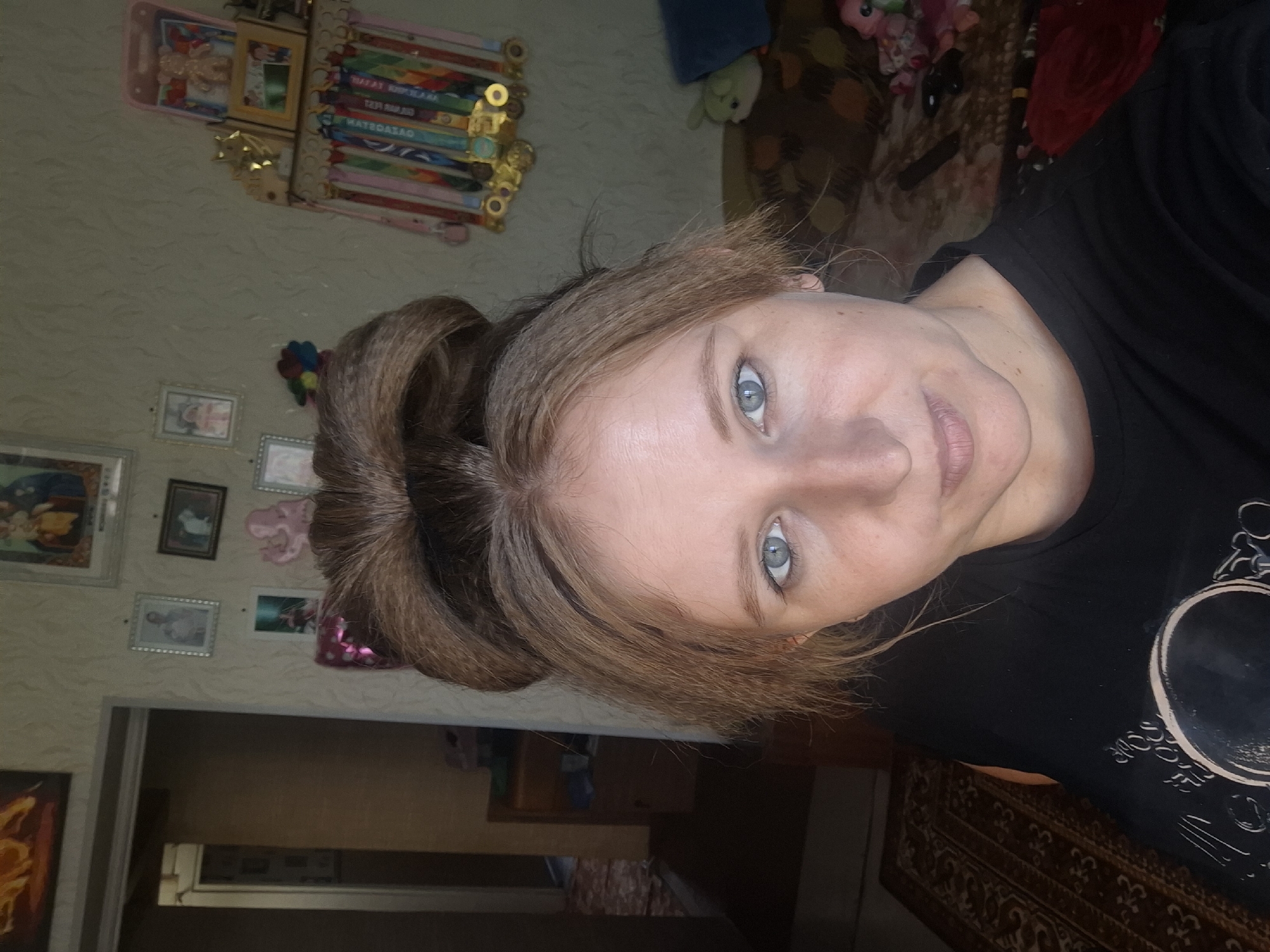 The image is a close-up, self-taken photograph of a woman with light brown hair styled in a large bun. She has blue eyes and fair skin. She is wearing a black t-shirt with a circular graphic on it. The background appears to be an indoor residential setting with patterned wallpaper. Several framed pictures are visible on the wall, along with a shelf displaying medals and small decorative items, including a pink teddy bear. To the left of the woman, a doorway leads into another room with a dark carpet. The scene is consistent with a domestic environment in Shymkent, Kazakhstan.