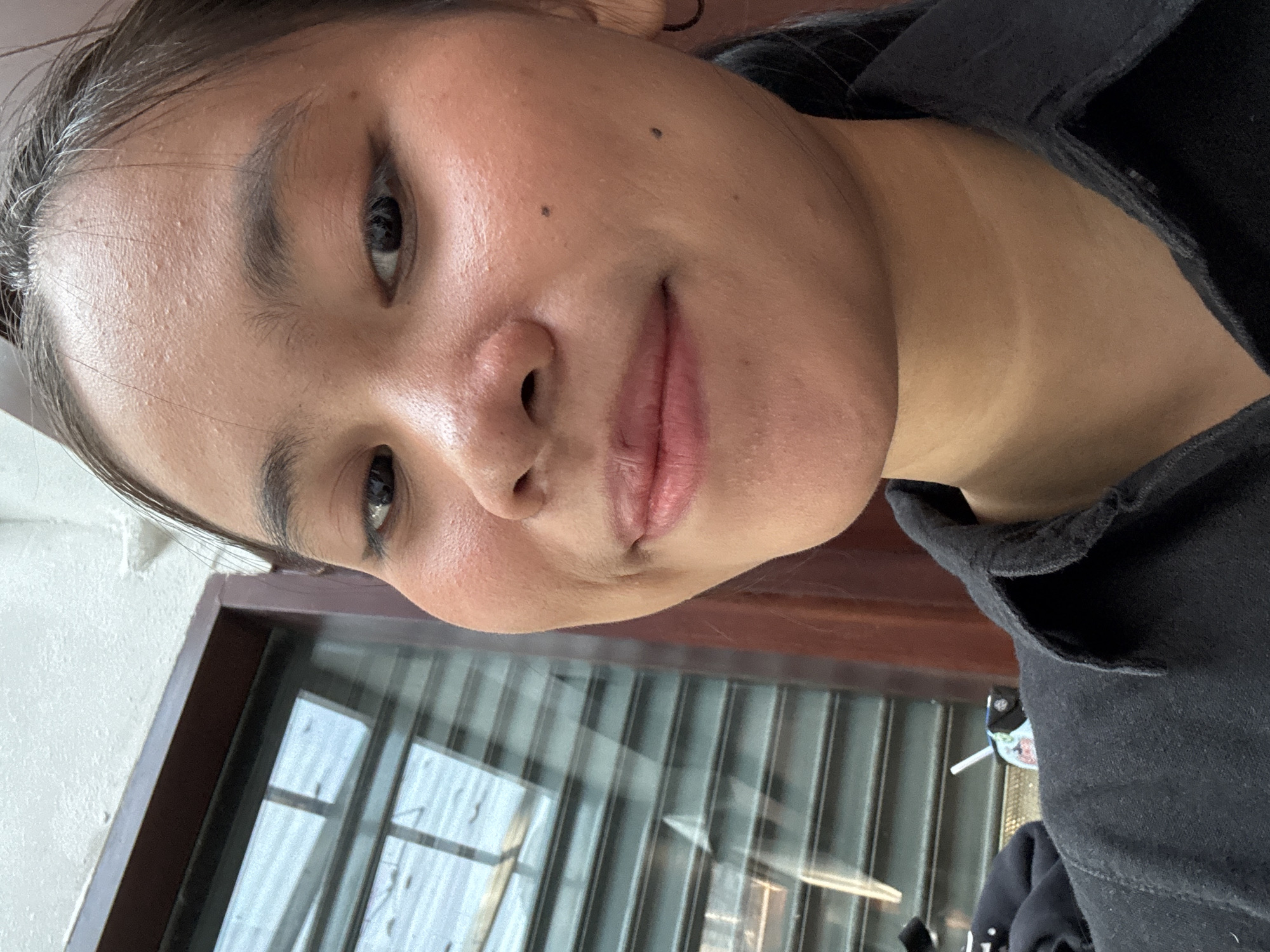 A medium shot captures a young woman from the chest up, facing the camera. She has dark hair pulled back and is wearing a black collared shirt. Her face is slightly turned, and she has a light smile. Her skin appears to have some texture and a few small blemishes on her forehead and cheek. She is wearing a subtle makeup. In the background, a window with horizontal blinds is visible to her left, and a dark wooden frame surrounds the window. The overall lighting suggests an indoor setting. The image is not specific enough to determine a location in South Jakarta, Indonesia.