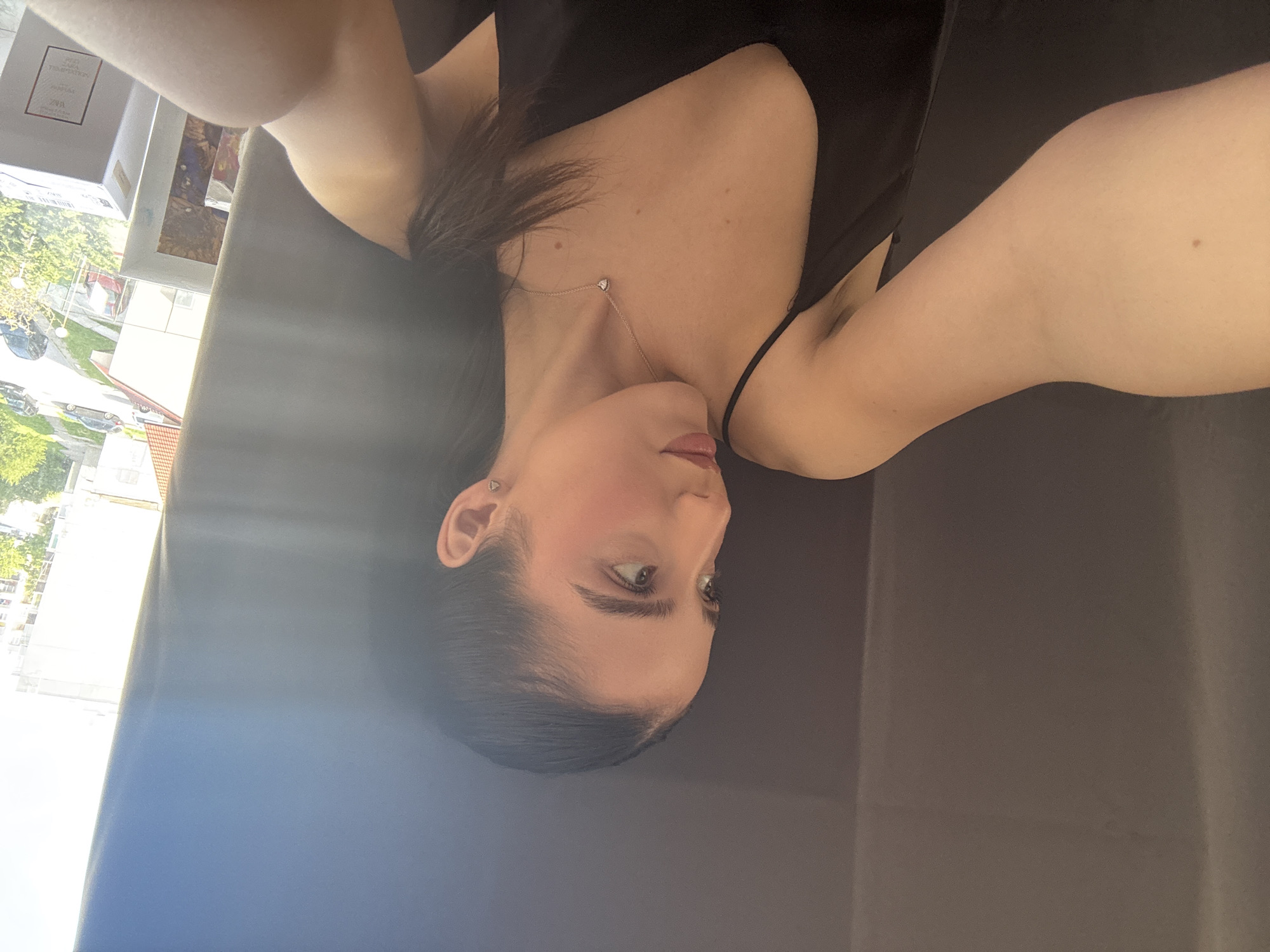 A woman with fair skin and dark hair is lying on a dark-colored surface, possibly a sofa or bed. She is wearing a black top and a delicate necklace with a heart-shaped pendant. The background shows a glimpse of an urban environment with buildings and trees, indicating a location within a city. The specific city or location within the city is unknown based on the image.