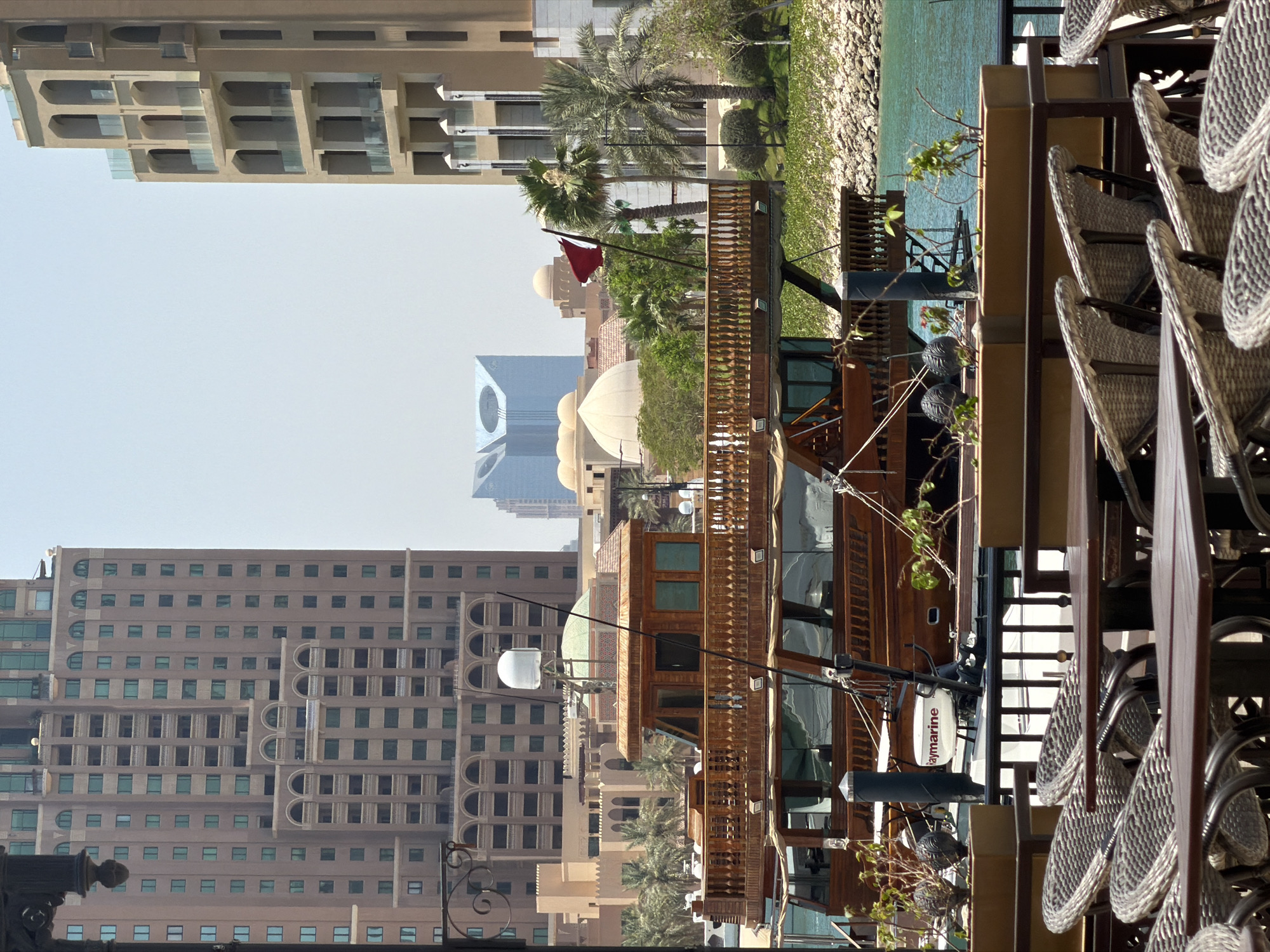 A waterfront scene in an urban setting features a multi-story wooden boat docked in turquoise water. The boat has several decks, intricate wooden railings, and large windows. Behind the boat, several tall buildings with beige and brown facades rise into a hazy sky. One building on the left is a large, rectangular structure with many windows and arched decorative elements. Another building on the right has a similar architectural style with arched openings and balconies. In the distance, a modern glass building with a distinctive geometric shape is visible. Palm trees and other greenery are scattered throughout the scene, with a rocky embankment visible along the edge of the water. In the foreground, an outdoor seating area with tables and wicker chairs is set up. A Qatari flag is prominently displayed on a pole near the boat. The overall impression is a sunny day in a resort-like environment.