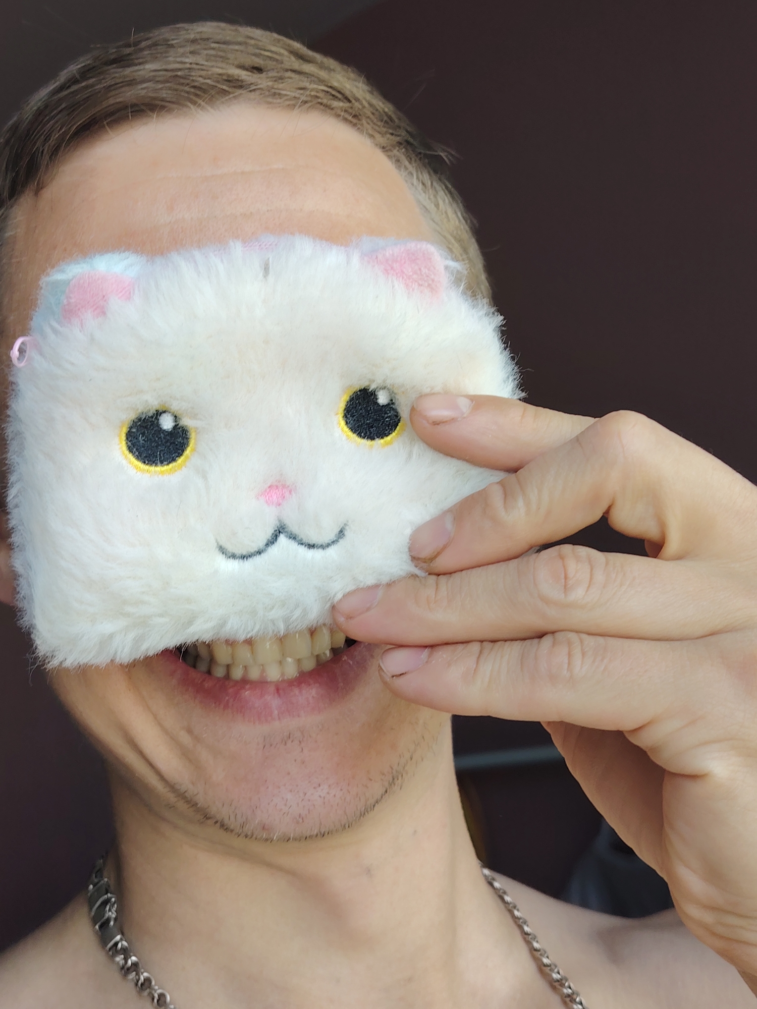 A person is smiling at the camera while holding a white, furry cat-shaped eye mask. The mask covers their eyes, and the person's hand is touching the mask. The person is wearing a silver chain necklace. The location is unknown, and there is no verifiable information to suggest this image was taken in Chaikovsky, Russia.