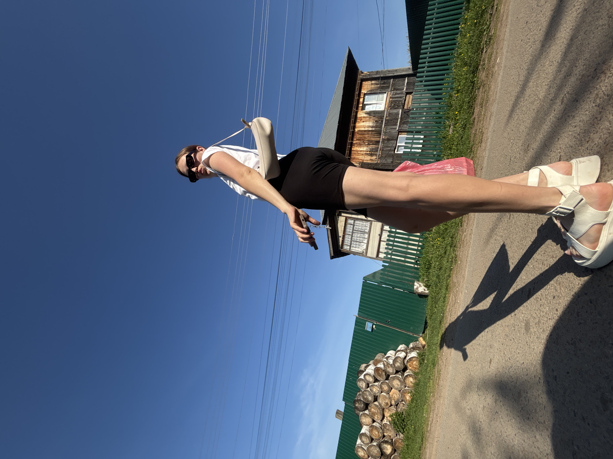A low-angle shot depicts a woman standing on a rural road in Фоки, Russia, against a clear blue sky. She is wearing a white top, black shorts, sunglasses, and white platform sandals. A small white purse hangs from her shoulder, and she holds a black object in her hand. In the background, a weathered wooden house sits behind a green metal fence. To the side, a pile of stacked logs rests on the grass. Power lines stretch across the sky above.