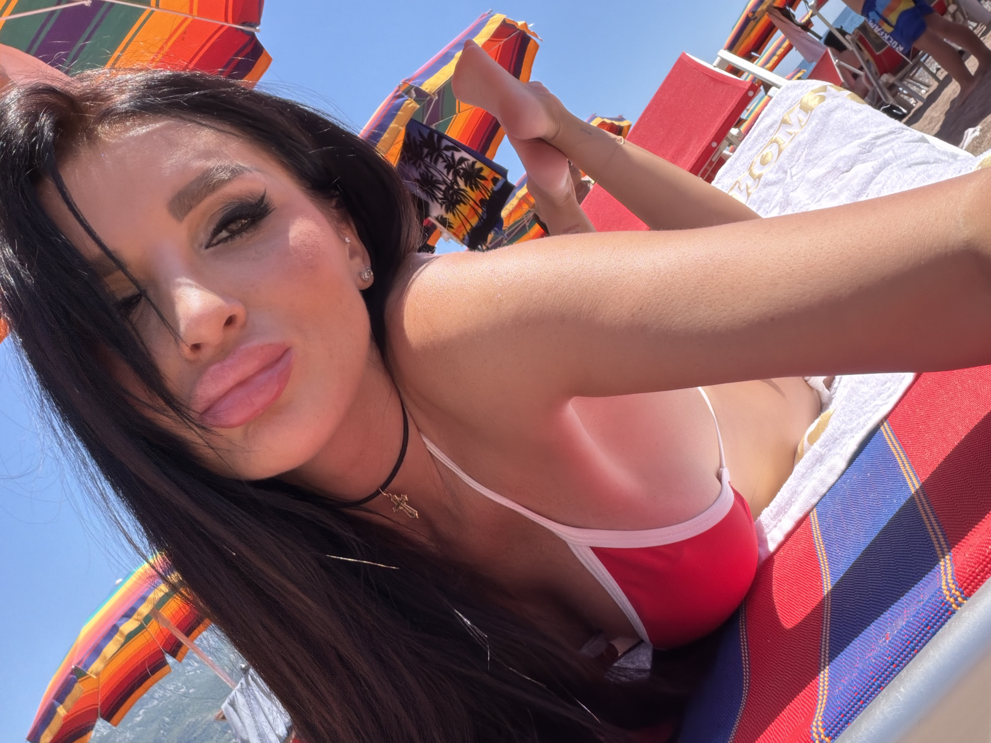 A young woman is depicted lying on a beach chair at a sunny beach in Boreti, Montenegro, taking a selfie. She has long, dark hair, prominent eye makeup, and full lips, which she is pouting with her tongue slightly visible. She is wearing a red bikini top with white trim, a black choker with a small golden cross pendant, and a stud earring. Her right arm and shoulder are visible in the foreground. The beach chair features red and blue striped fabric, and a white towel with a partial gold inscription (possibly "HOME") is draped over it. In the background, under a clear blue sky, numerous colorful striped beach umbrellas are visible, some with patterned designs including palm trees. The sea is partially visible on the left side, and other red beach chairs with vague figures of other individuals are present in the distance.