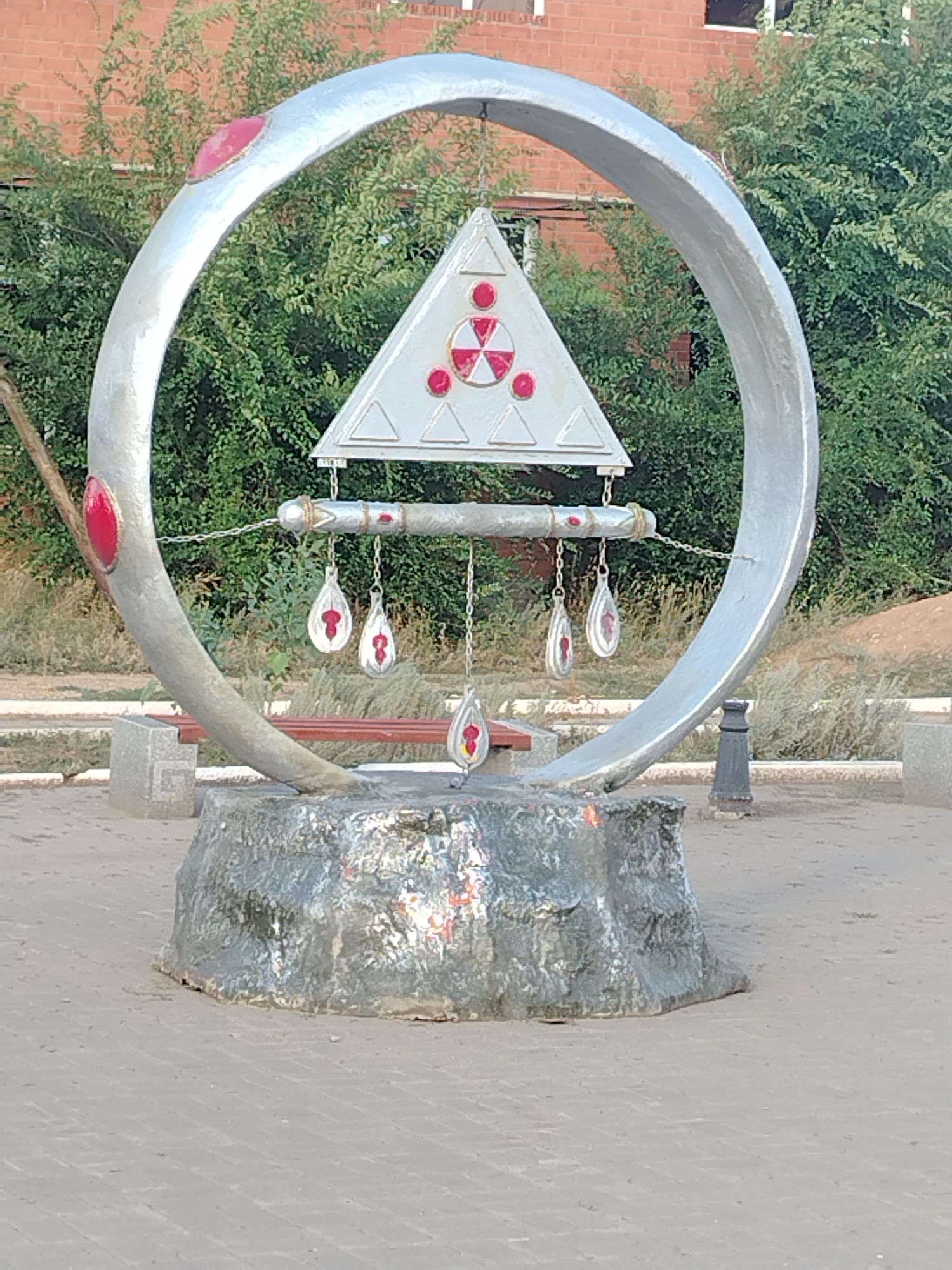 The image shows an outdoor monument located in the city of Temirbek Jürgenov village, Kazakhstan. The monument is a circular metal structure, painted silver, with red circular accents. Suspended from a horizontal bar within the circle is a triangular metal plate adorned with a symbol resembling a radiation hazard sign, also featuring red circular accents. Below this, several teardrop-shaped metallic ornaments hang, each with red interior detailing. The monument is mounted on a textured, rock-like base, which appears to be made of concrete or a similar material. In the background, there is dense green foliage and a brick building. The ground in front of the monument is paved with light-colored rectangular stones. There are no people visible in the image.
