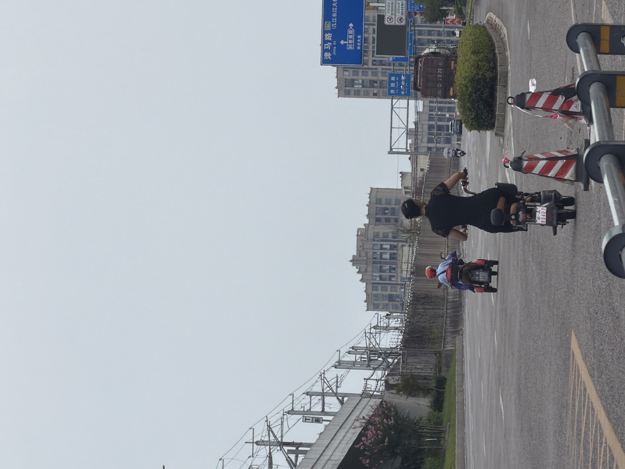 The image depicts a street scene in Jiangjin, China, under an overcast sky. In the foreground, two people are riding electric scooters. The scooter on the right is ridden by a woman wearing a black dress with lace sleeves. The scooter on the left is ridden by a person wearing a blue shirt and a red helmet, with a red backpack. In the background, there are several buildings, a highway overpass with train tracks, and traffic signs. One sign indicates directions to "津马路 (Jiuma Rd)" and "科教南路 (Kejiao South Rd)". There are also traffic cones on the road, suggesting roadwork or a traffic diversion. The overall environment appears to be a busy urban area with infrastructure related to transportation.