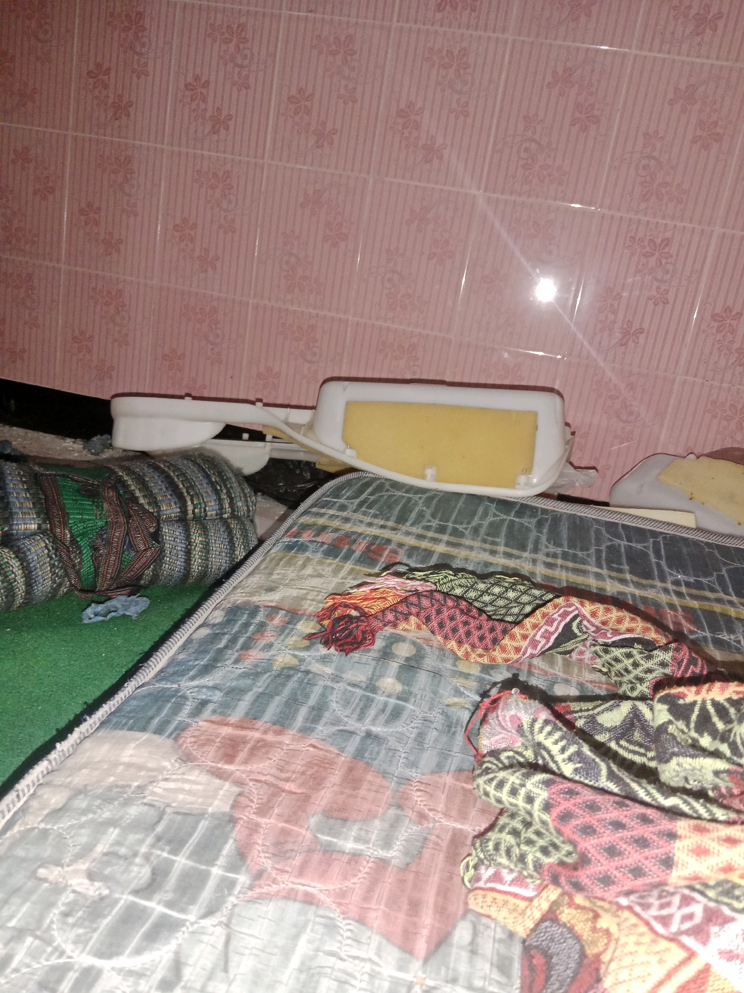 The image depicts a cluttered interior scene with a pink, floral-patterned tiled wall in the background. In the foreground, a mattress is visible, covered with a patterned quilt. Several fabric items, including a green mat and a rolled-up item with green and blue stripes, are placed on or near the mattress. A white plastic object with a yellow foam insert rests on top of some of the fabric items. There are no people visible in the image. The scene appears to be in a domestic setting, possibly a bedroom or storage area. There is no specific information about the location in Ringinrejo, Indonesia, provided in the image itself.