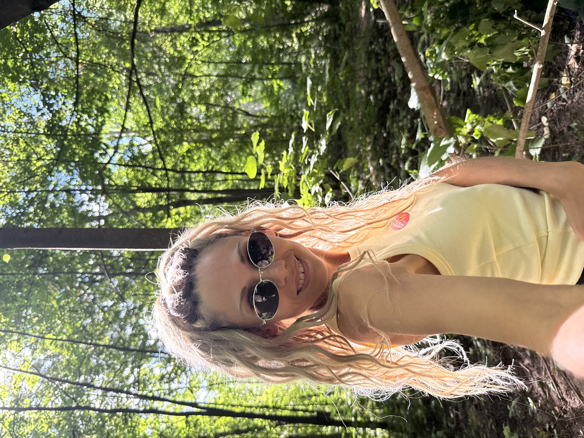 In a sun-dappled forest, a woman with blonde, curly hair tied into a high ponytail poses for a selfie. She wears oval, silver-rimmed sunglasses and a light yellow tank top with a pink circular logo on the left chest. The background is filled with lush green trees and foliage, with sunlight filtering through the leaves, creating a bokeh effect. The ground is covered in leaf litter and fallen branches, suggesting a natural, wooded environment. The woman is smiling and looking directly at the camera, her right arm extended holding the phone. The location is identified as Rusanovo, Russia.