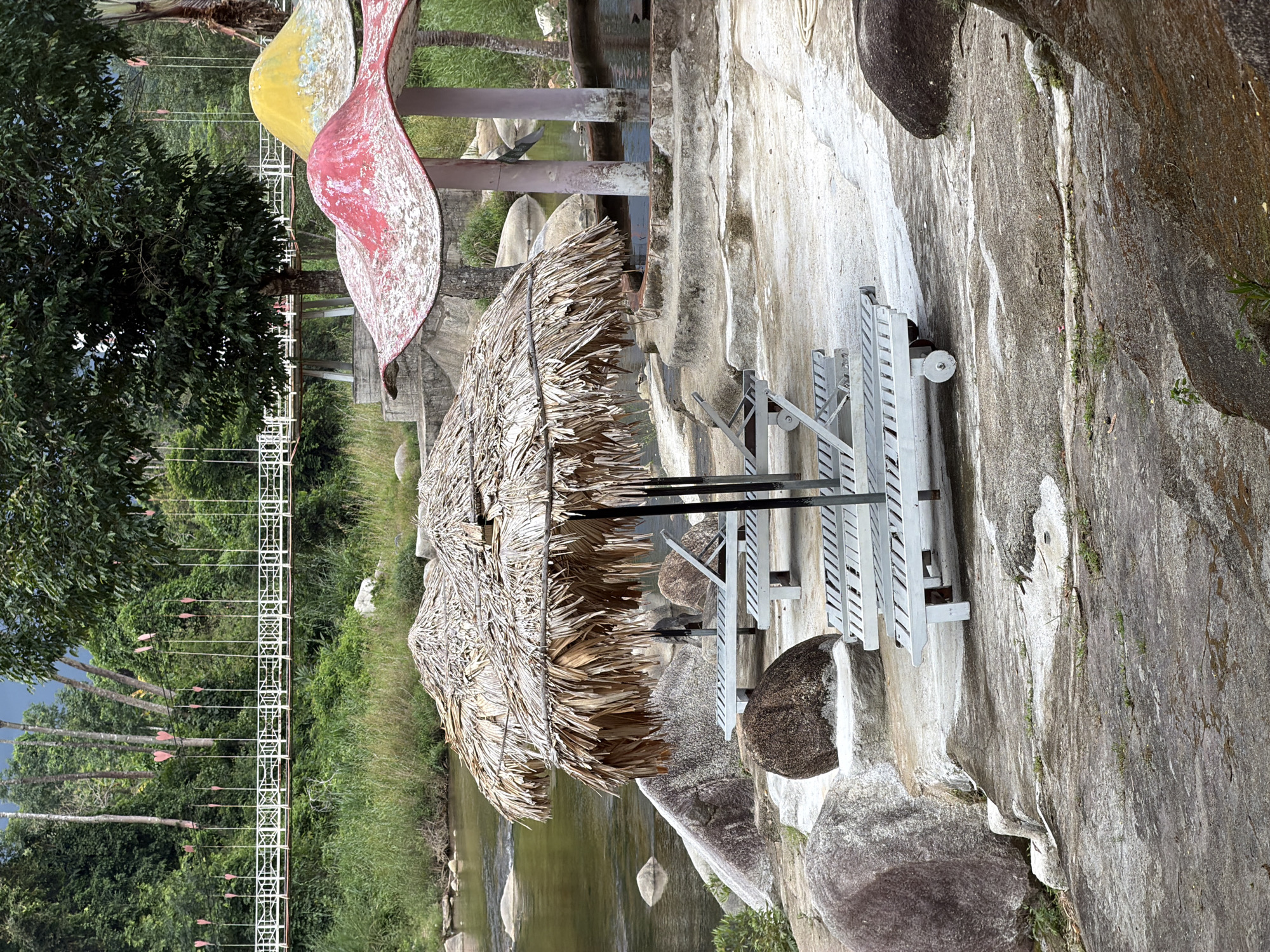 The image depicts a riverside resort area in Khanh Vinh, Vietnam, featuring a tranquil natural setting. In the foreground, two thatched-roof umbrellas cast shadows over several white lounge chairs situated on a concrete platform adjacent to a rocky riverbank. The river itself, a calm body of water with scattered rocks, flows gently to the left of the frame. Behind the lounge chairs, a series of curved, mushroom-shaped structures, one predominantly red and the other yellow, provide additional shade. These structures appear to be part of a decorative water feature or seating area. In the background, a suspension bridge with white latticework spans across the river, adorned with numerous small pink heart-shaped decorations. Lush green foliage and tall trees dominate the landscape, creating a dense forest backdrop. The sky above is overcast, suggesting a humid or potentially rainy day. The overall impression is one of a peaceful, rustic getaway designed for relaxation and enjoying the natural environment.