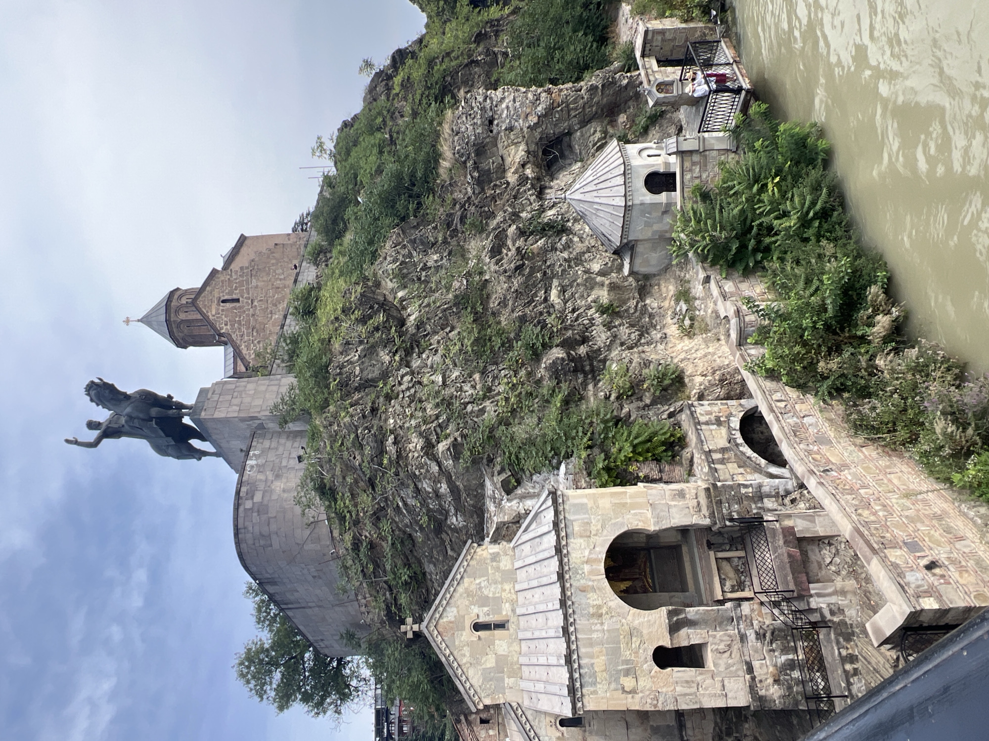 The image shows a view of the Narikala Fortress and the Holy Trinity Cathedral of Tbilisi, Georgia, with the Kura River in the foreground. At the top of the image, a large equestrian statue of King Vakhtang Gorgasali stands prominently on a hilltop overlooking the city. To the right of the statue, the dome and bell tower of the Holy Trinity Cathedral are visible. Below the fortress and cathedral, on the rocky hillside, are several smaller churches and structures. In the lower left of the image, a stone church with a red-tiled roof and an arched entrance is visible. Further to the right, along the riverbank, is a small gazebo-like structure and a viewing platform where two individuals, one wearing a white hat and the other a red dress, are standing. The Kura River, appearing muddy, flows along the bottom of the frame. The architecture suggests a historical and religious site, typical of Tbilisi. The day appears overcast with clouds in the sky.