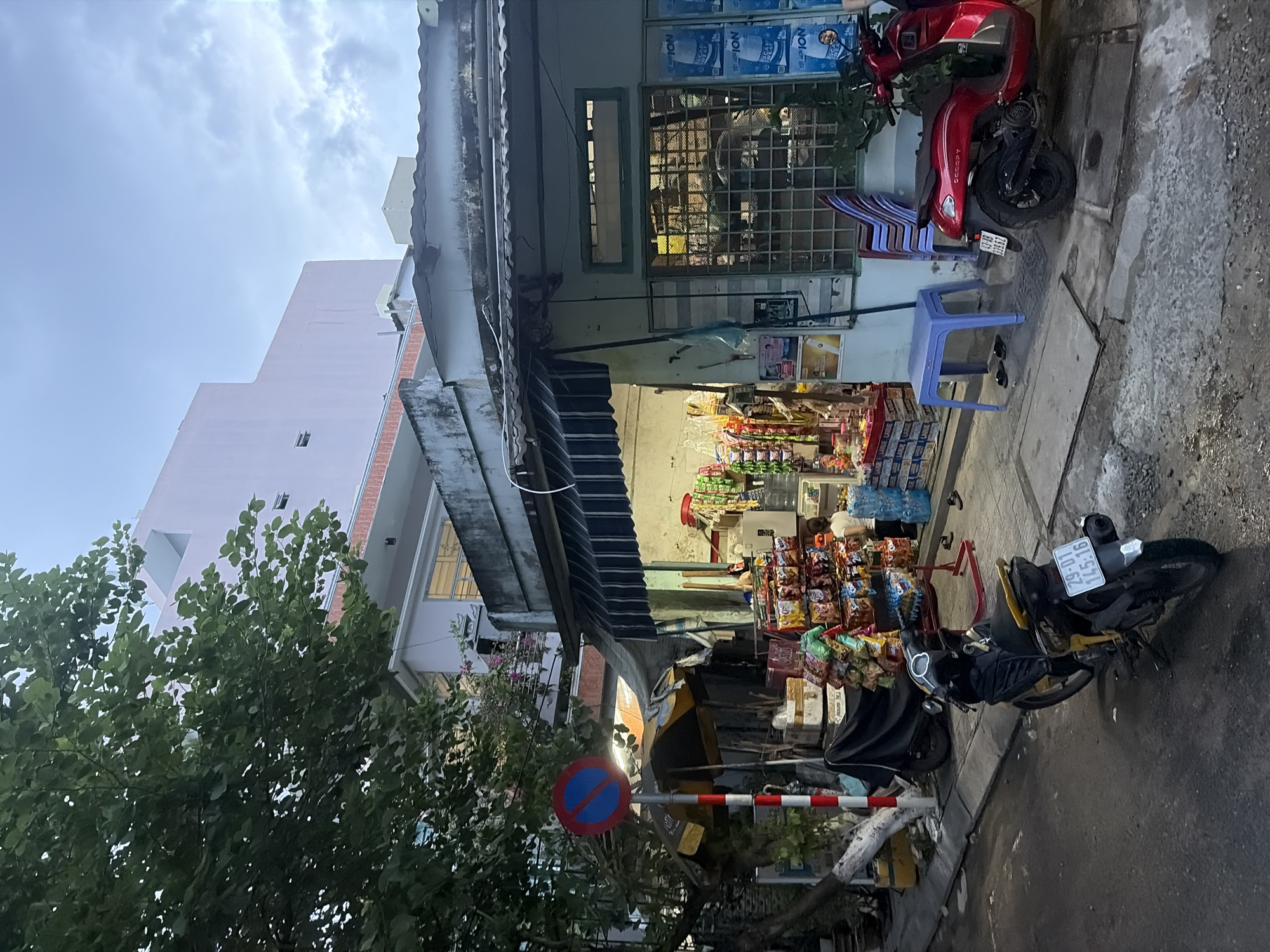 The image depicts a street scene in Da Nang, Vietnam, likely in the evening or twilight hours, judging by the artificial lighting and the sky. A small convenience store is visible on the right, with its interior lit and stocked with various goods, including snacks and bottled drinks displayed on shelves. Two motorcycles are parked on the street in front of the store; one is black and yellow, and the other is bright red. A person, appearing to be a woman, is standing inside the store. A blue plastic stool and some chairs are placed outside the store. A traffic sign indicating "No Stopping" is visible on the left side of the street, next to a tree and a yellow and black umbrella. In the background, buildings of different heights and architectural styles are present, suggesting a mixed urban environment. The street surface is paved with asphalt and concrete.